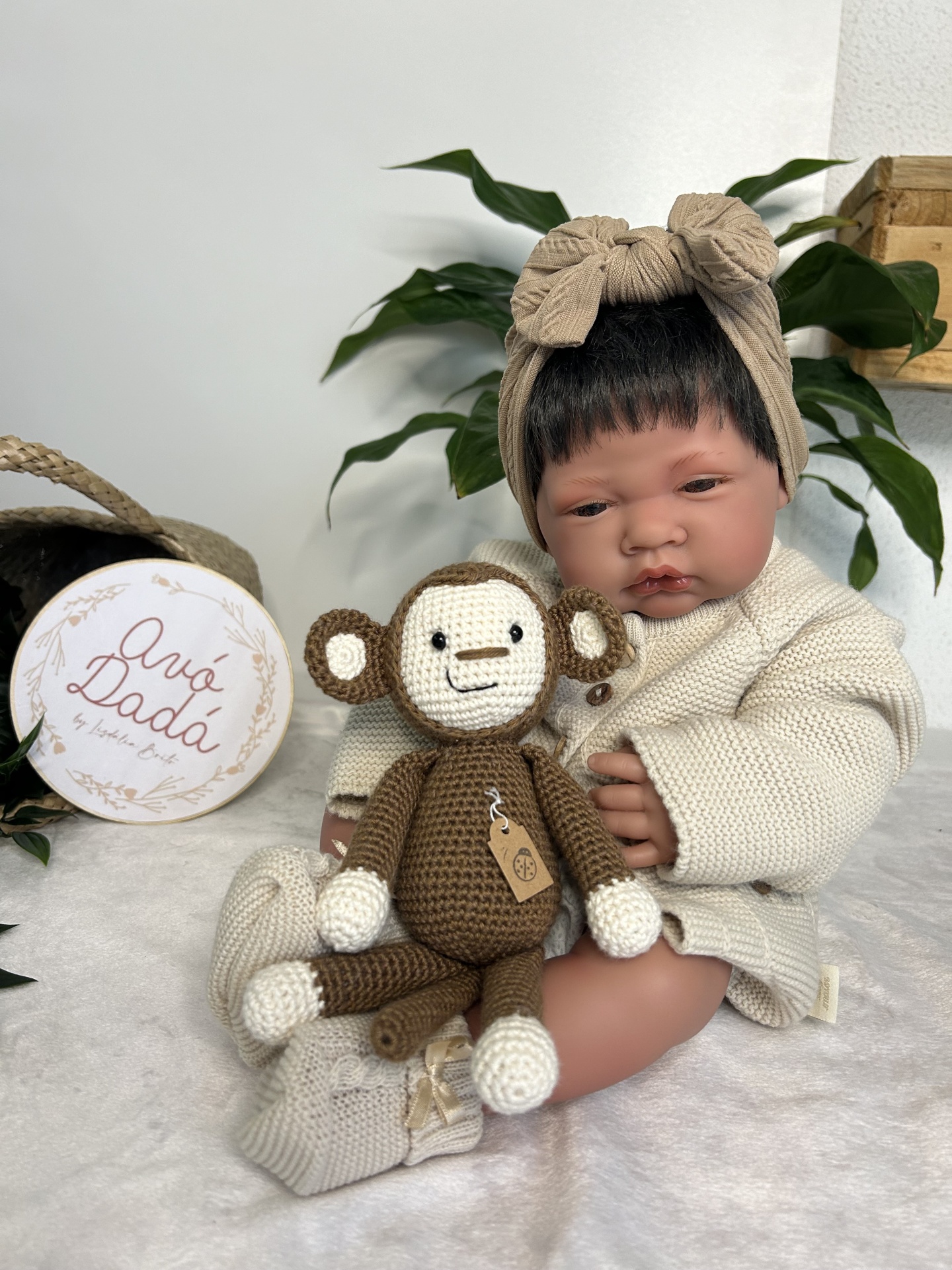 Boneca bebé com roupa de malha e faixa na cabeça, segurando macaco de croché, planta e caixa ao fundo