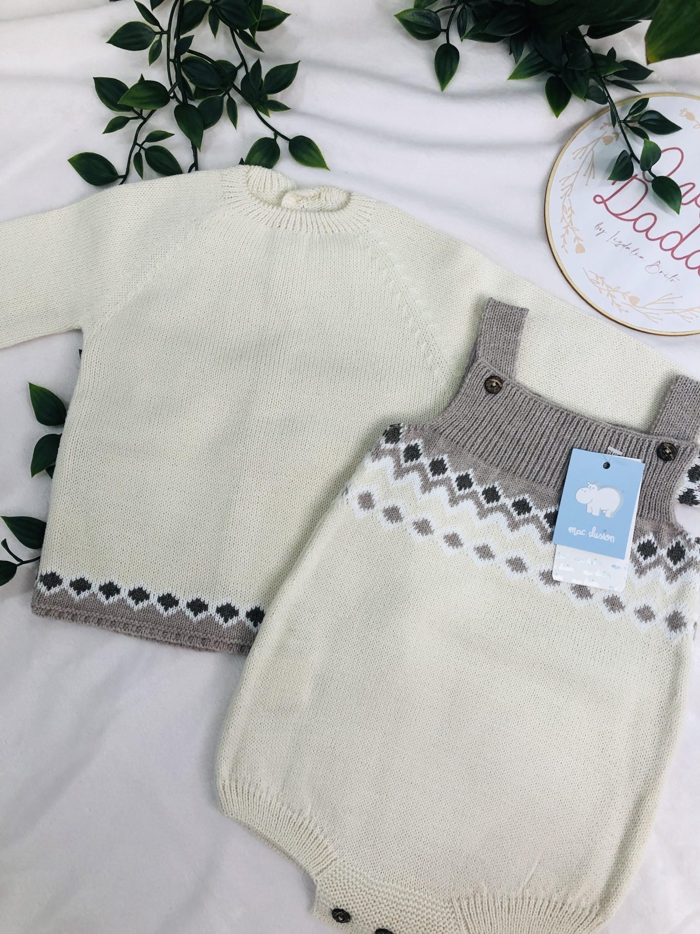 Conjunto de roupa de bebé em malha branca com padrão cinzento e bege, incluindo camisola e jardineiras, sobre tecido branco com folhas verdes.