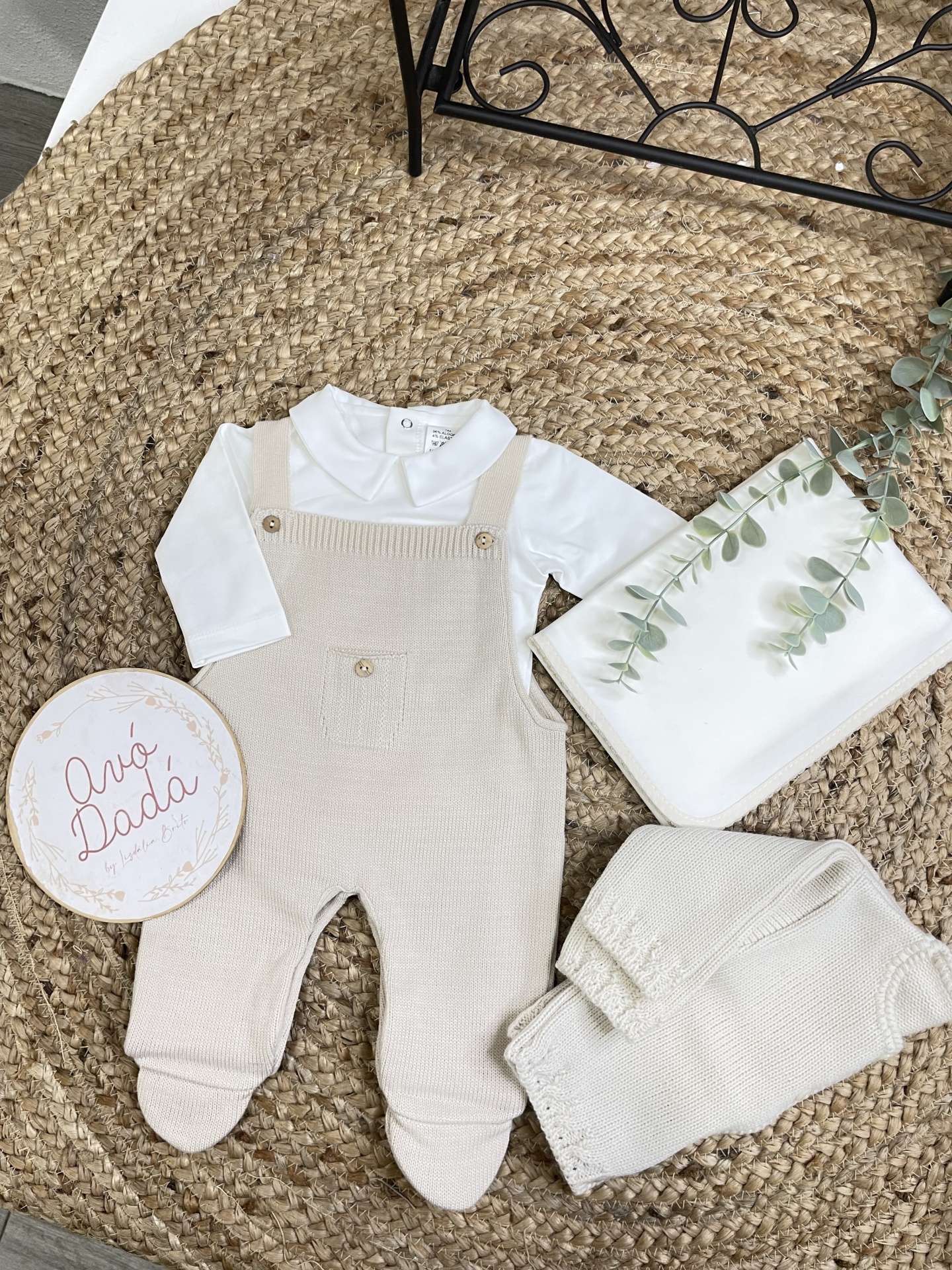 Conjunto de roupa de bebé com body branco e jardineira de malha bege sobre tapete de fibras naturais
