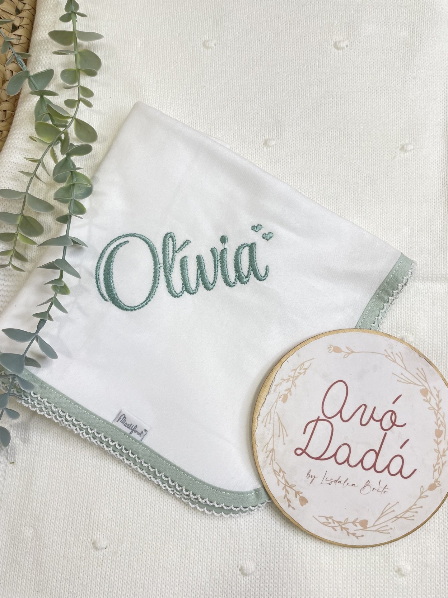Manta branca bordada com nome Olívia e detalhe verde claro com placa redonda decorativa ao lado