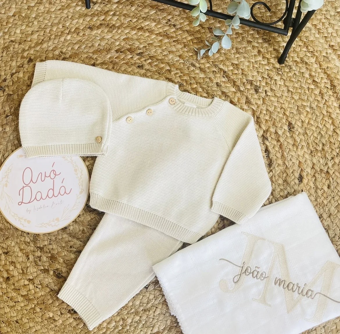Conjunto de roupa de bebé creme com manta branca bordada e placa decorativa num tapete de ráfia.