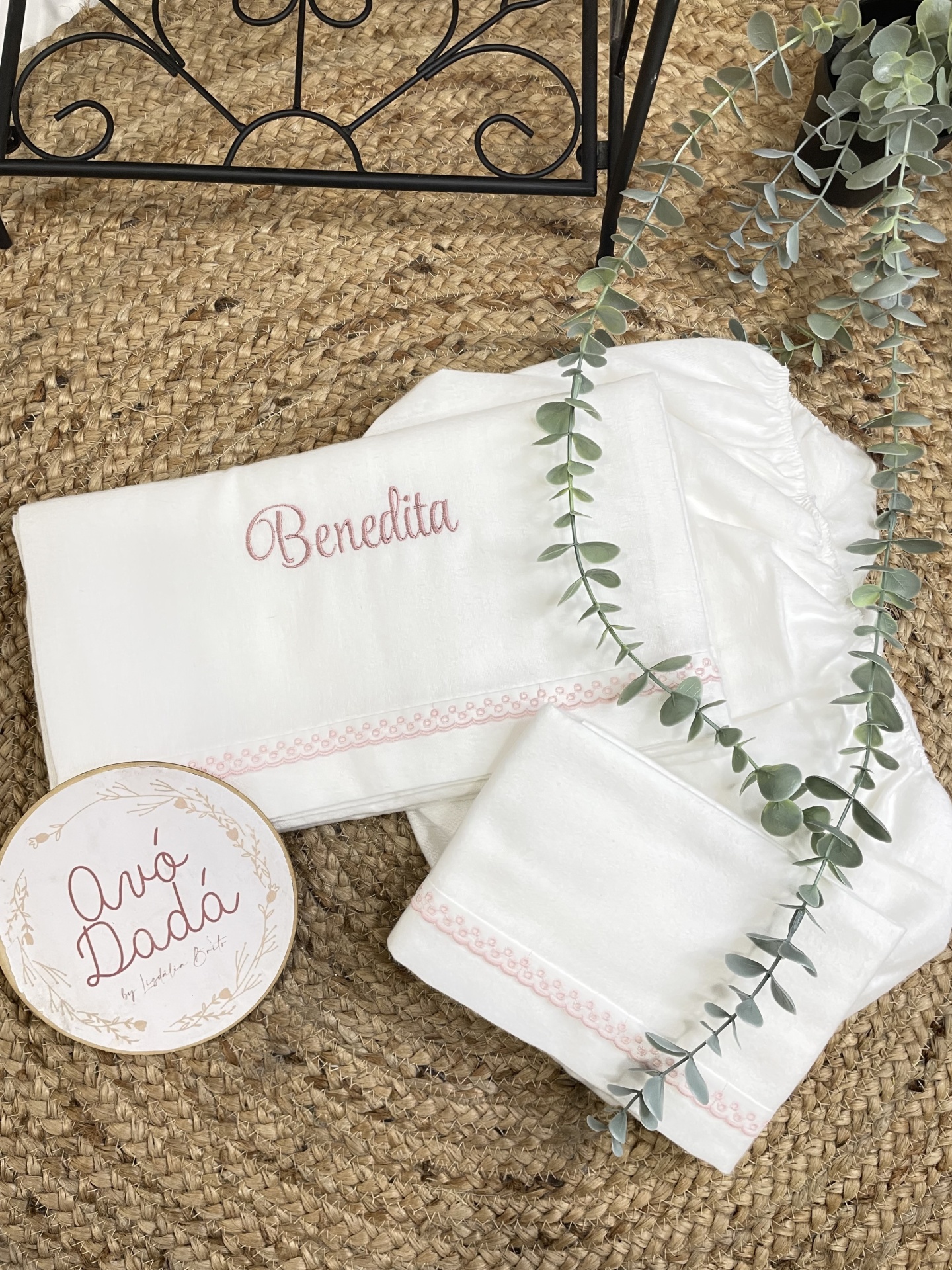 Conjunto branco de roupa de bebé bordado com o nome Benedita, sobre tapete de palha rústico.
