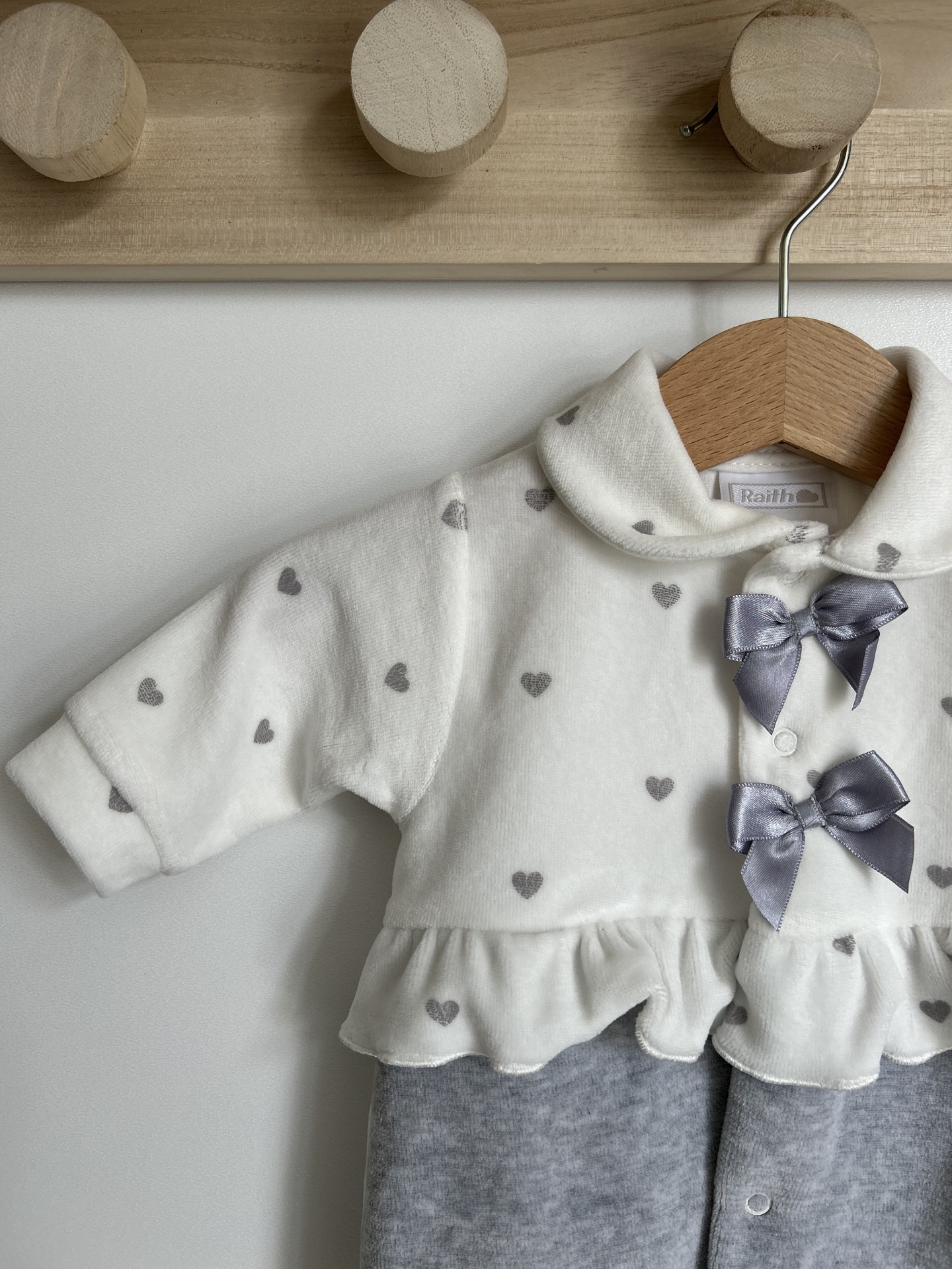 Macacão de bebé branco com corações cinzentos, laços cinzentos e parte inferior cinzenta pendurado em cabide de madeira