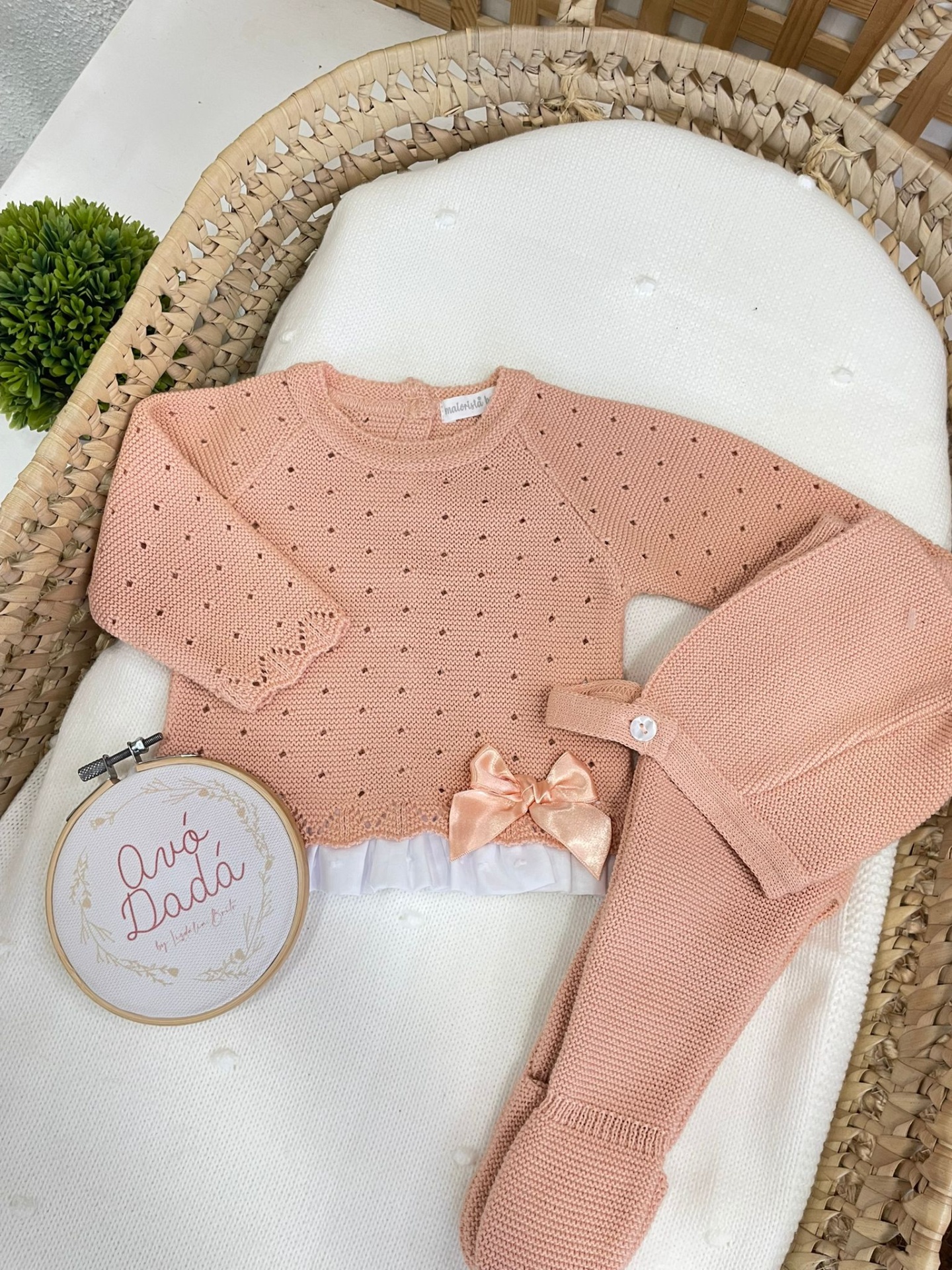 Conjunto de bebé rosa claro em malha com laço e saia branca sobre almofada branca em cesta de vime