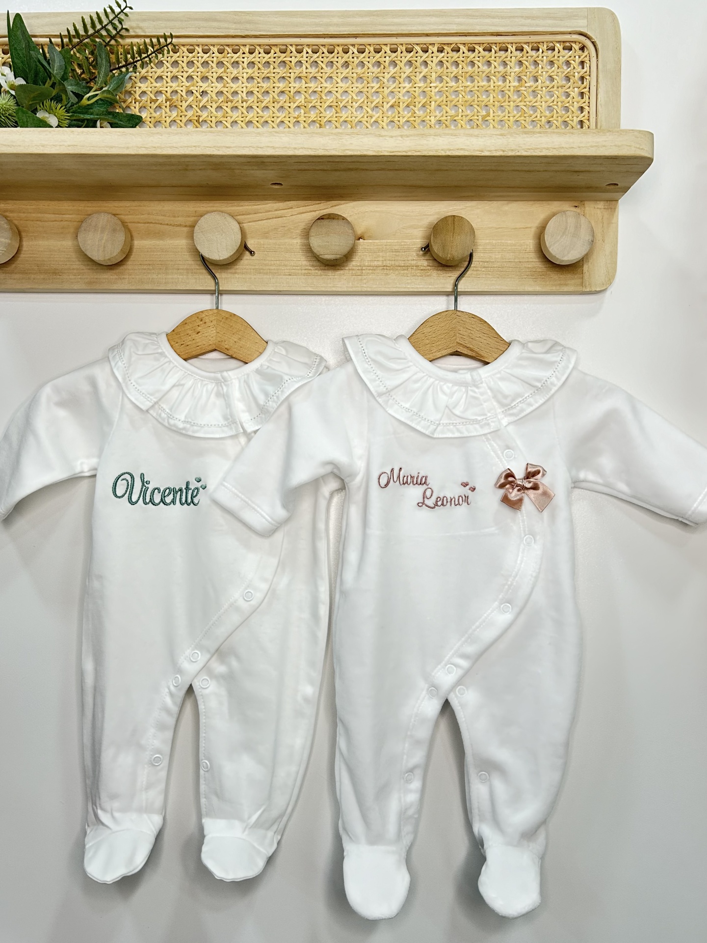 Dois macacões brancos para bebé com nomes bordados pendurados em cabides de madeira num cabide de parede
