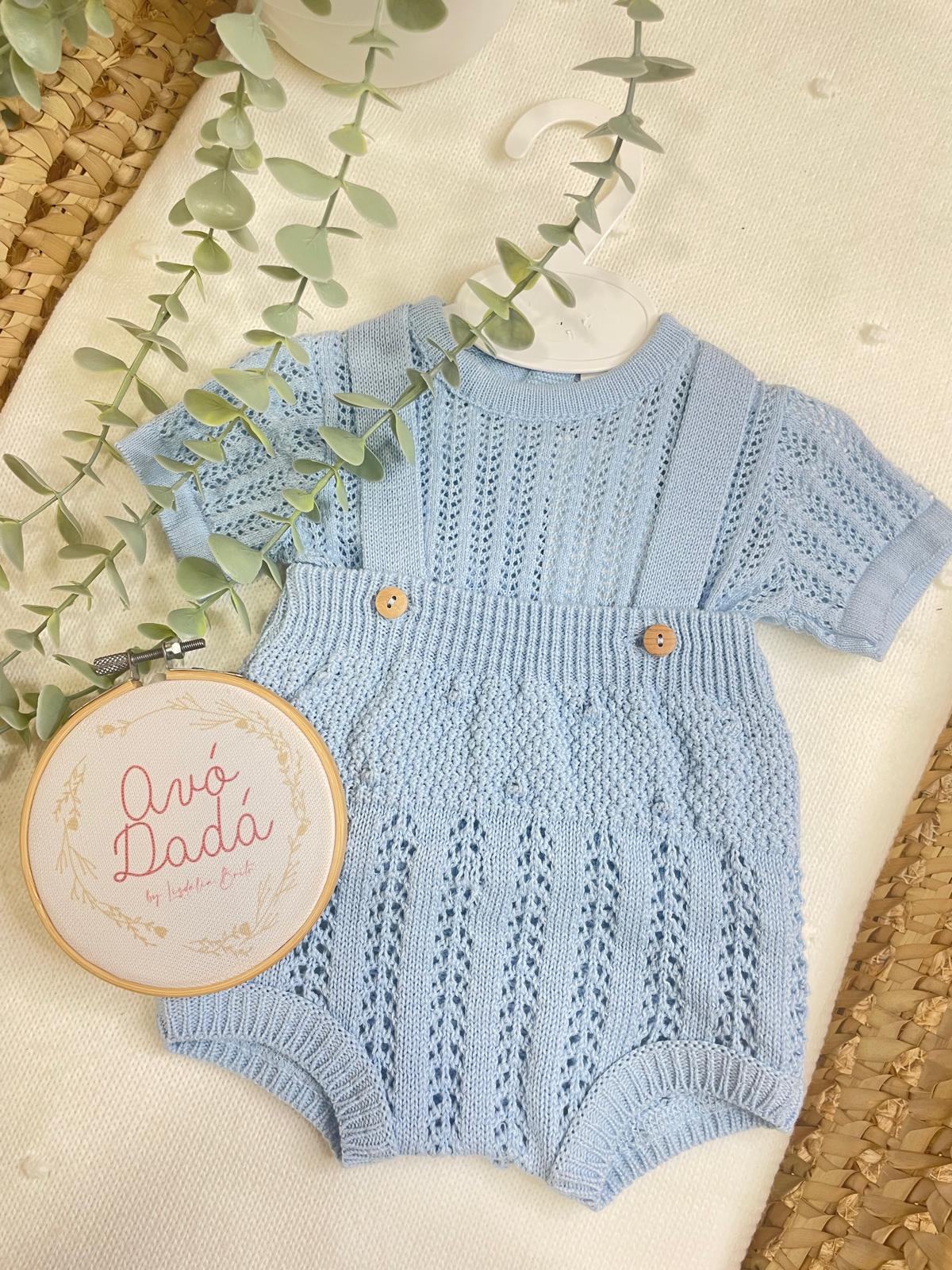 Conjunto de roupa de bebé azul clara em malha com padrão, com alças e botões de madeira sobre tecido claro e plantas decorativas.