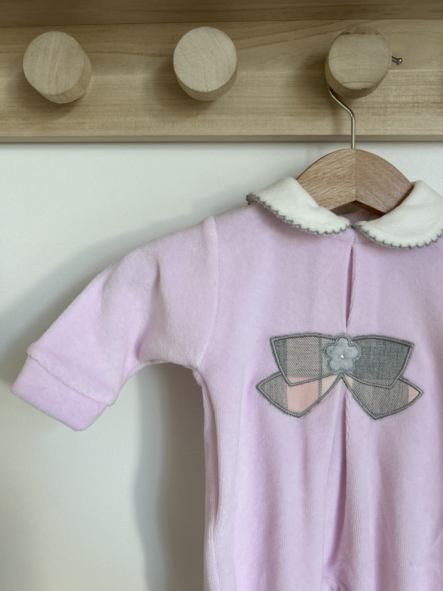 Macacão bebé rosa claro com gola branca e laço decorativo cinza e rosa, pendurado em cabide de madeira