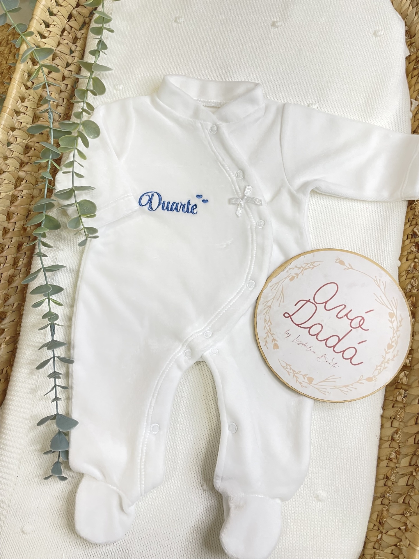 Macacão branco para bebé com bordado azul e laço branco dentro de cesto de palha com manta branca e folhas verdes