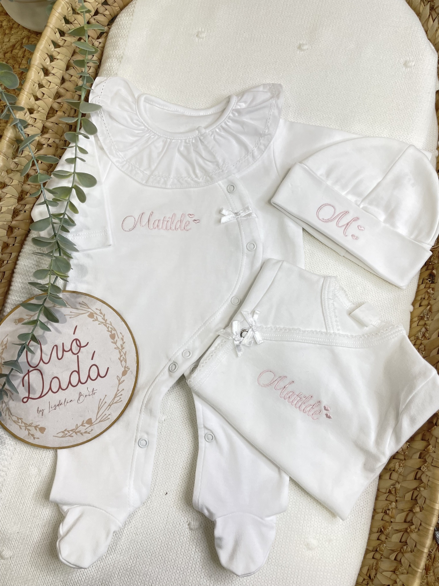 Conjunto de roupa branco de bebé com bordados cor-de-rosa Matilde e M:, com folho e laços, exposto em almofada e cesta de palha.