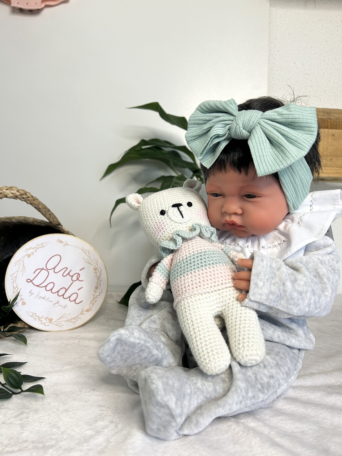 Boneca bebé realista com fato de pijama cinzento e fita de cabelo verde água segurando urso de tricot