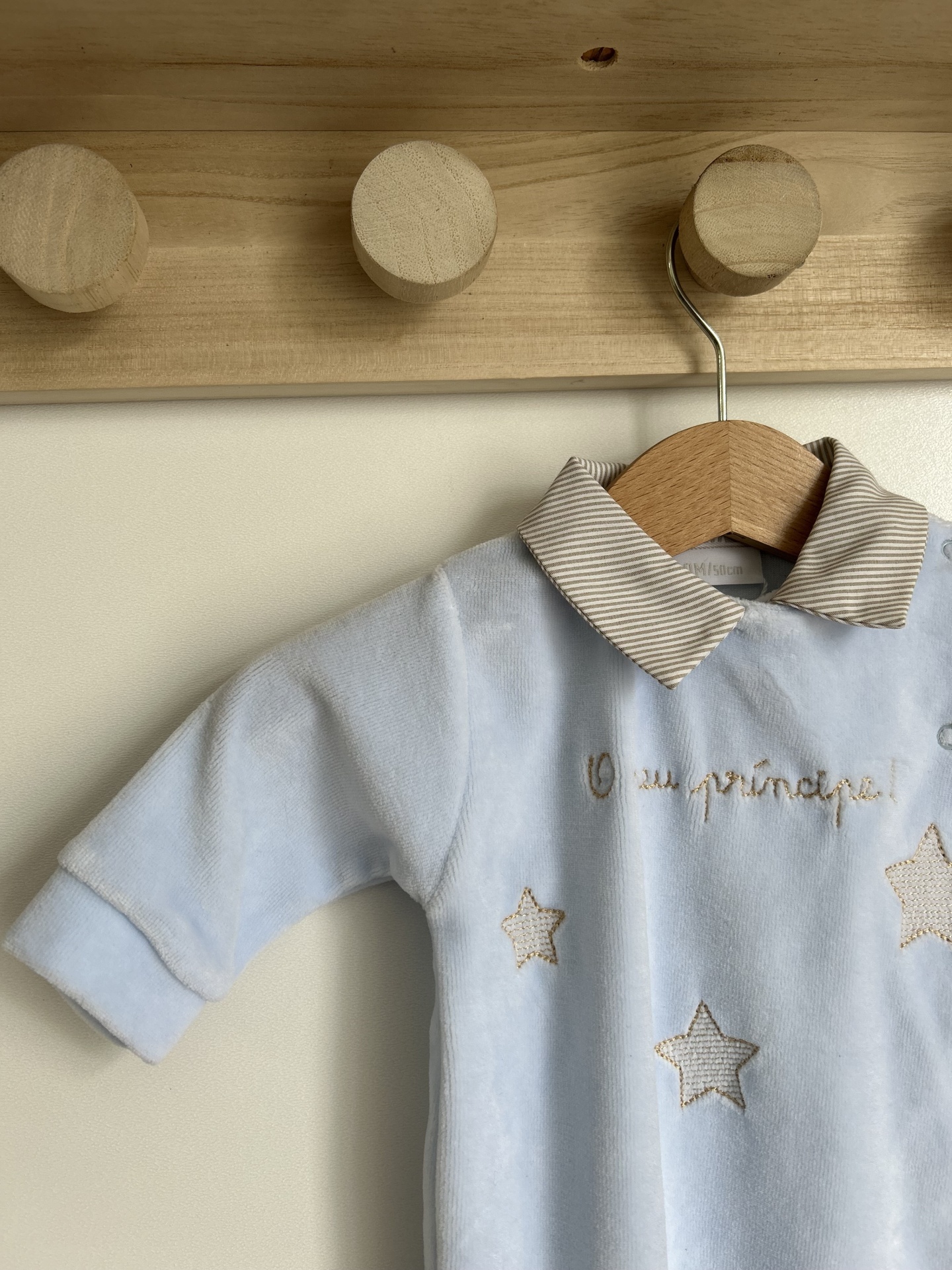 Pelele azul claro com gola riscada bege e branco e estrelas bordadas pendurado em cabide de madeira