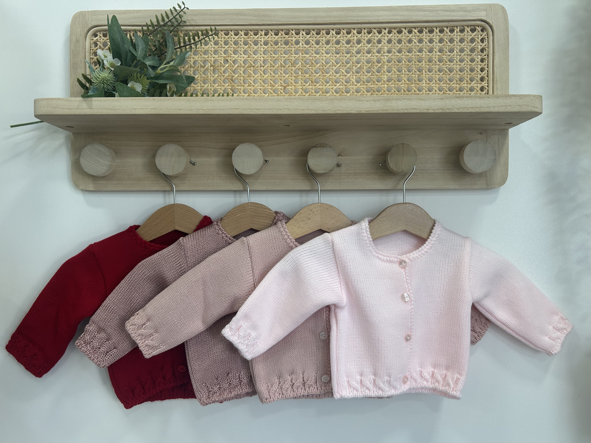 Conjunto de casacos de malha para bebé pendurados numa prateleira de madeira com vime e planta decorativa