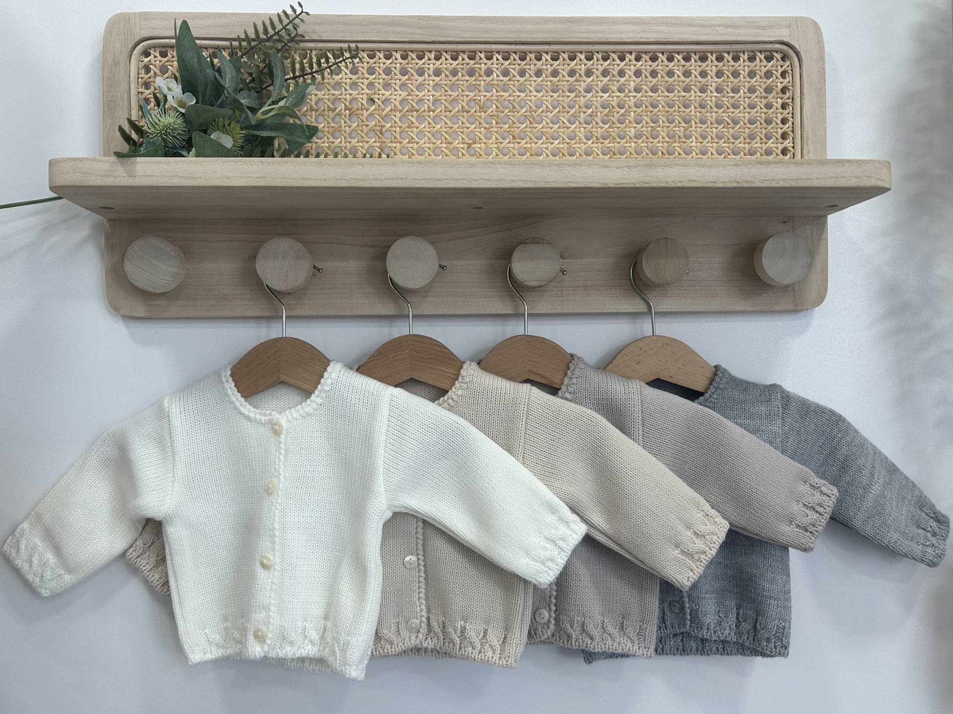 Quatro casacos infantis de malha em tons neutros pendurados em cabides de madeira sob prateleira em madeira com detalhe em palha.