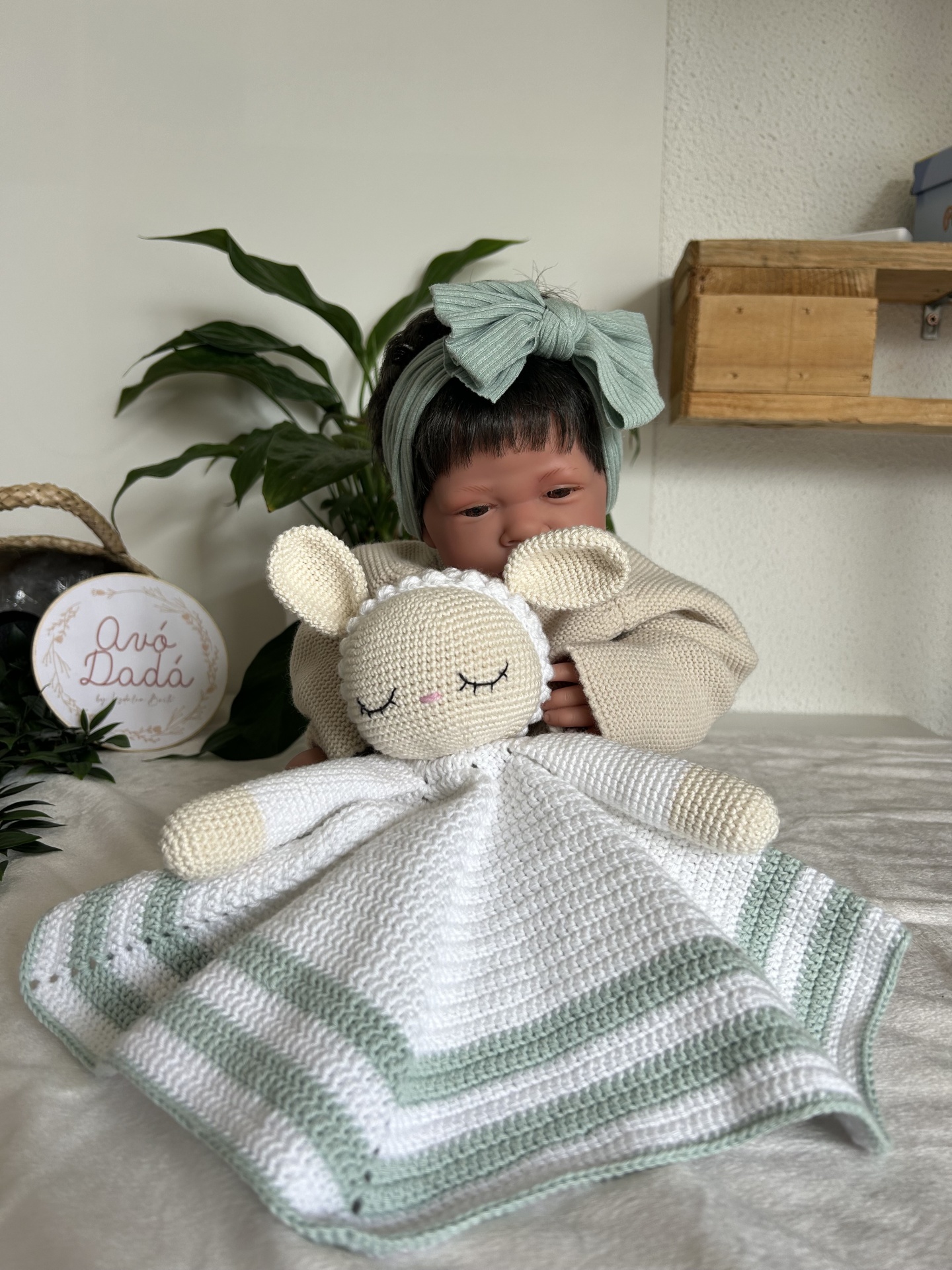 Boneca bebé a segurar cobertor de crochê com ovelha, em cenário com planta e prateleira de madeira