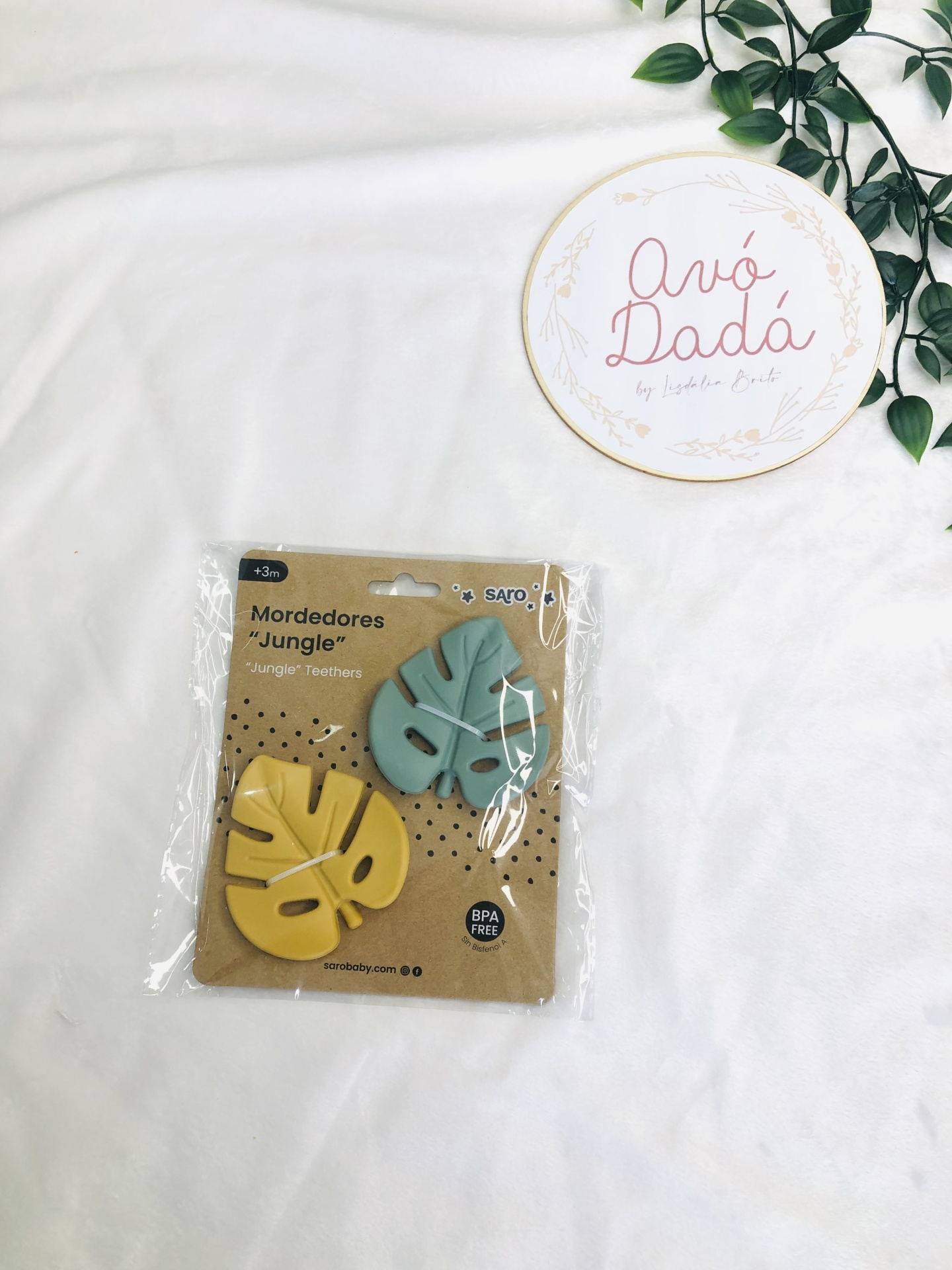 Dois mordedores em forma de folha amarelo e verde em embalagem, sobre tecido branco com raminho verde e círculo decorativo com texto Avó Dadá.