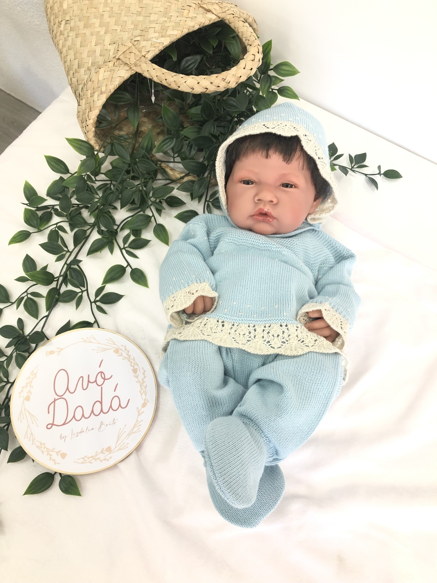Boneca realista vestida com roupa azul clara e capuz, com placa branca e cesta de palha ao lado.