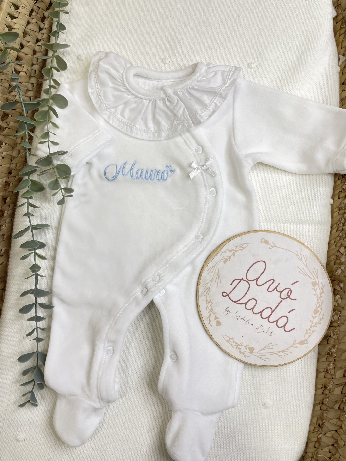 Macacão branco para bebé com gola franzida e bordado azul Mauro, em tecido branco e cesto de palha com planta verde e placa redonda