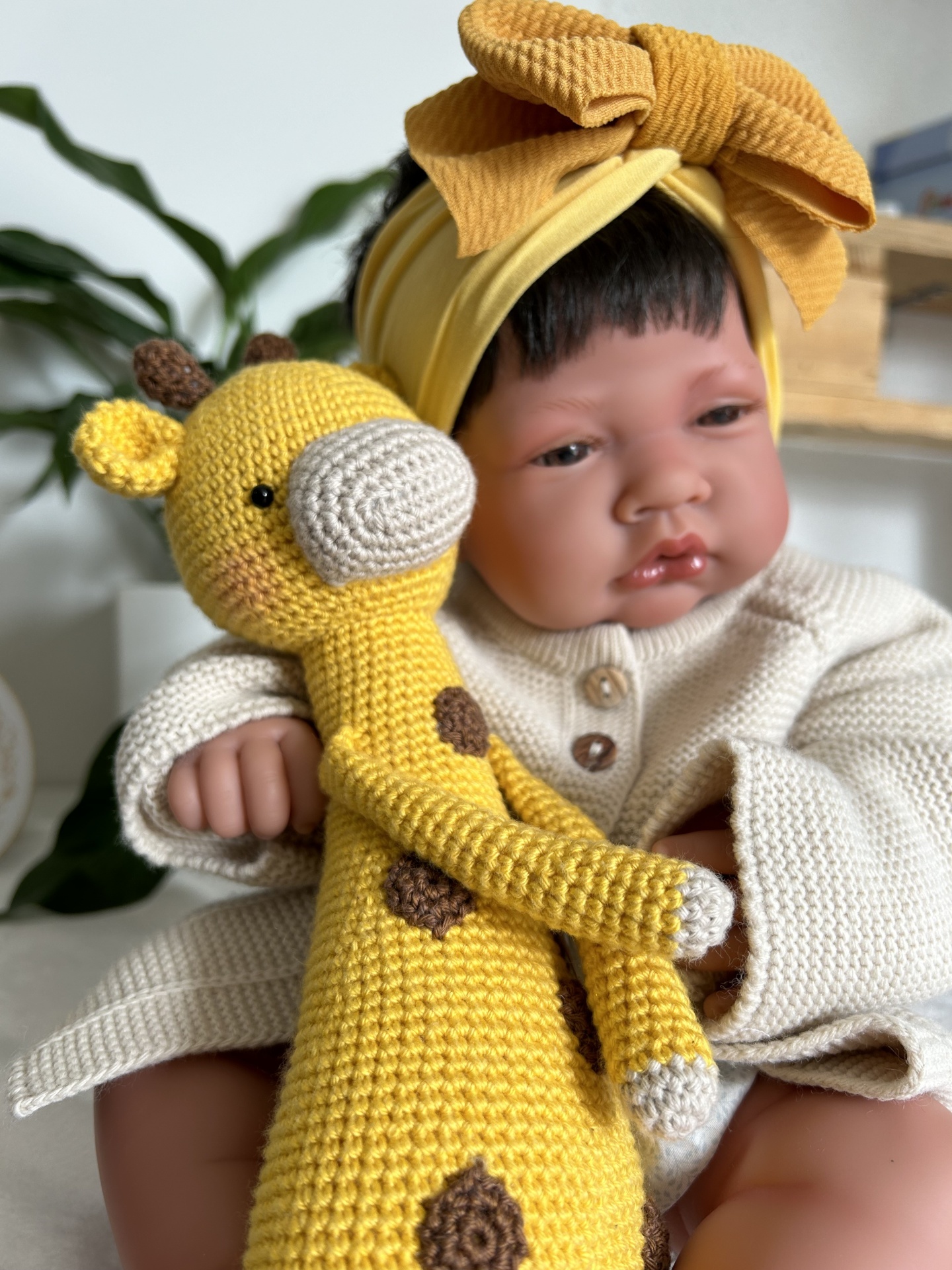 Boneca bebé com roupinha creme e touca amarela segurando giraffa de croché