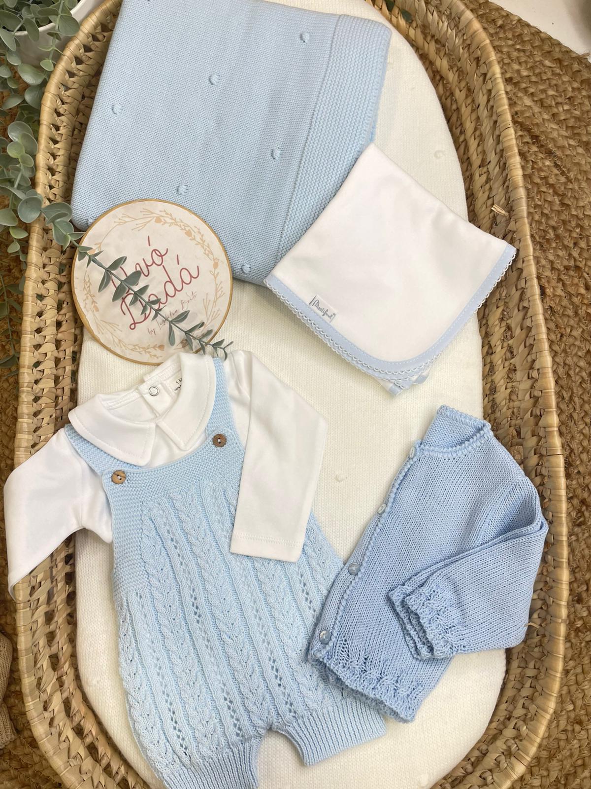 Conjunto de roupa azul clara de malha para bebé com body branco e acessórios numa cesta de verga.