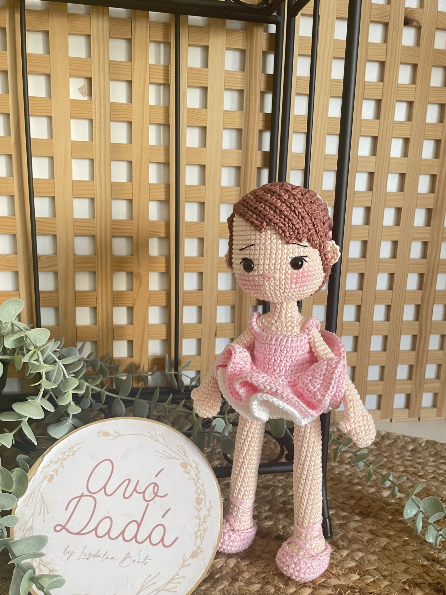 Boneca de croché com vestido rosa sentada com placa branca e planta ao fundo.