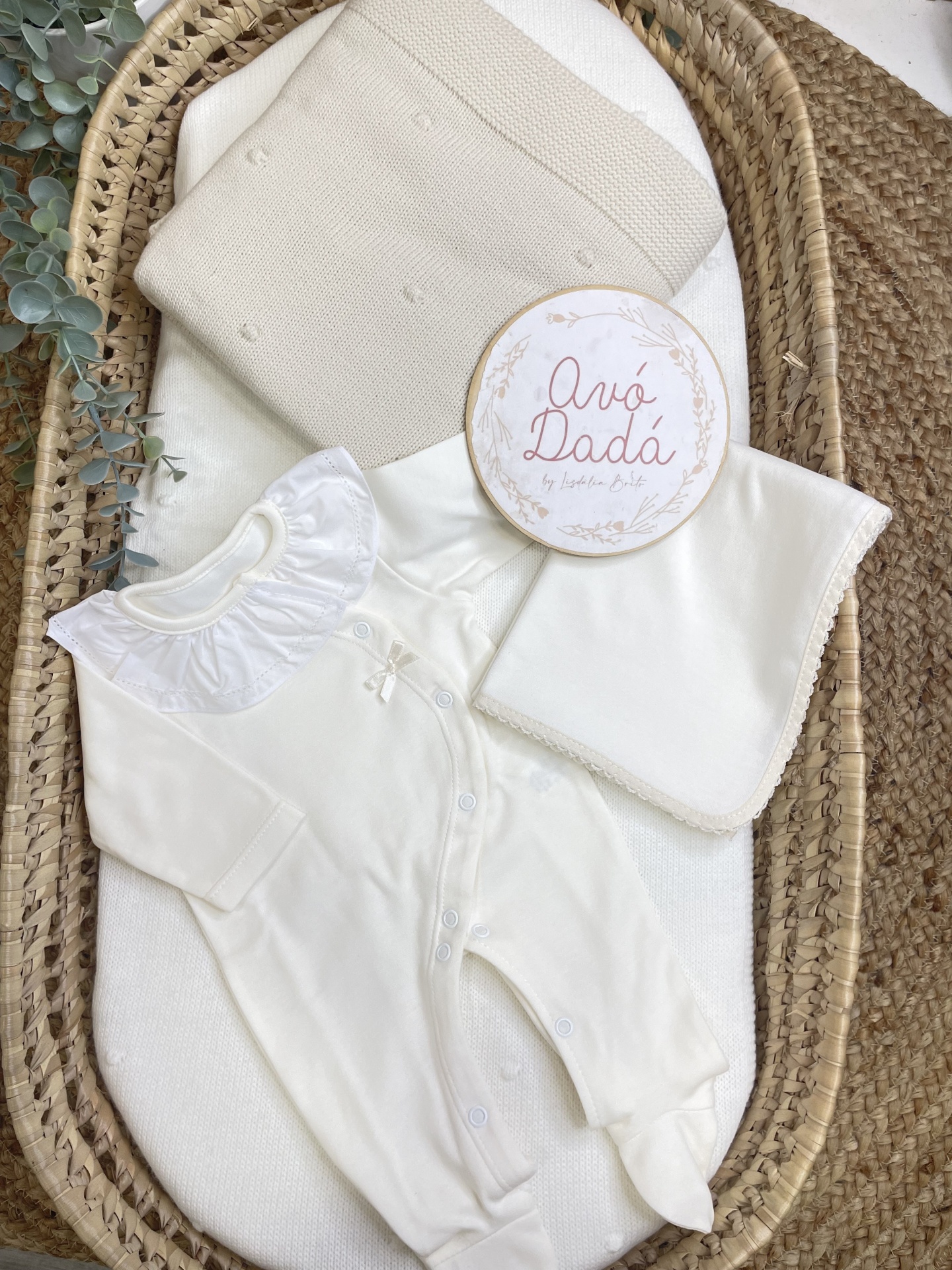 Conjunto de bebé branco com manta bege e babado no colarinho numa cesta de rattan.