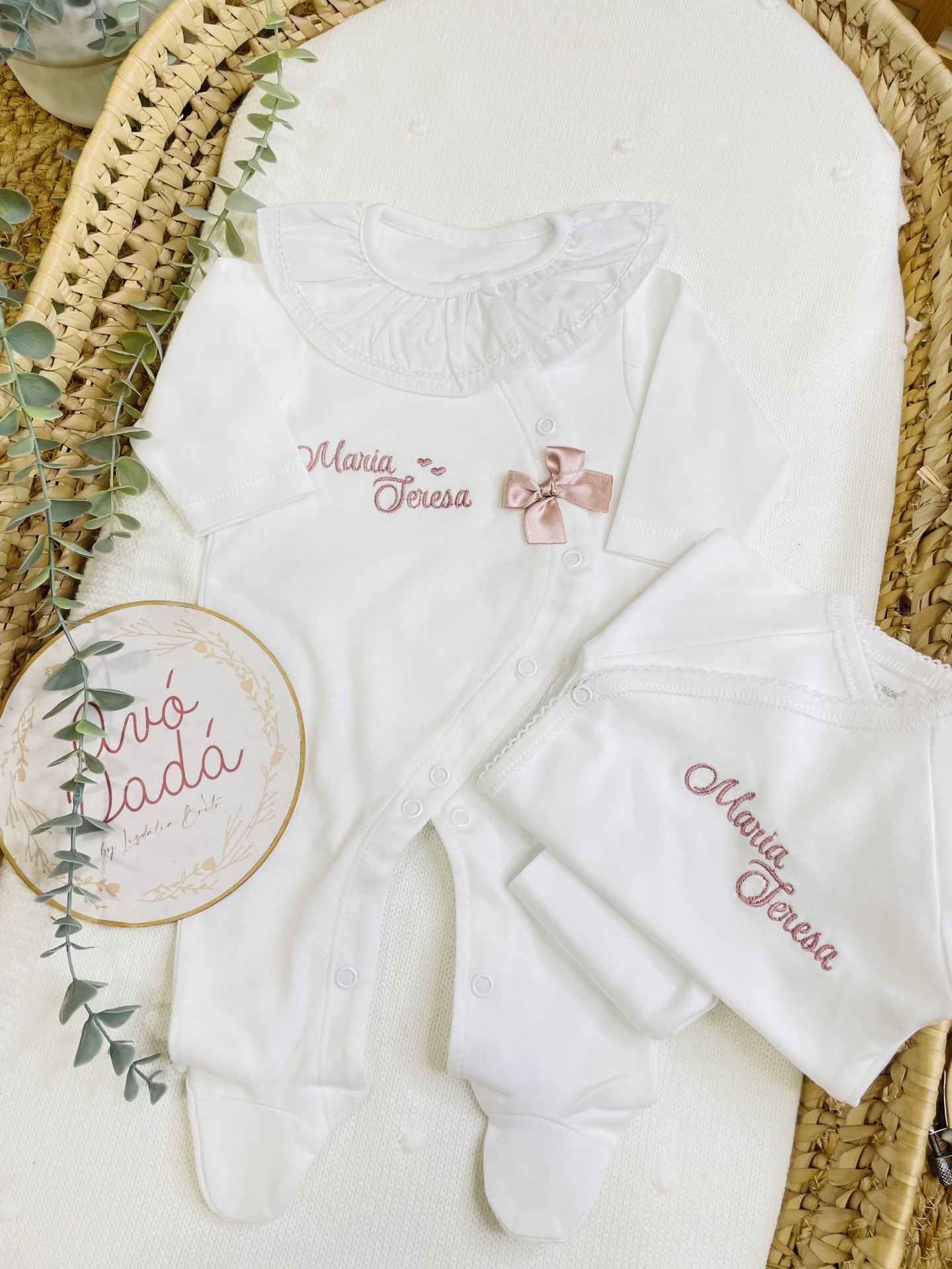 Conjunto de roupa de bebé branco com bordado rosa Maria Teresa e toalha, sobre colchão branco e cesta de vime.
