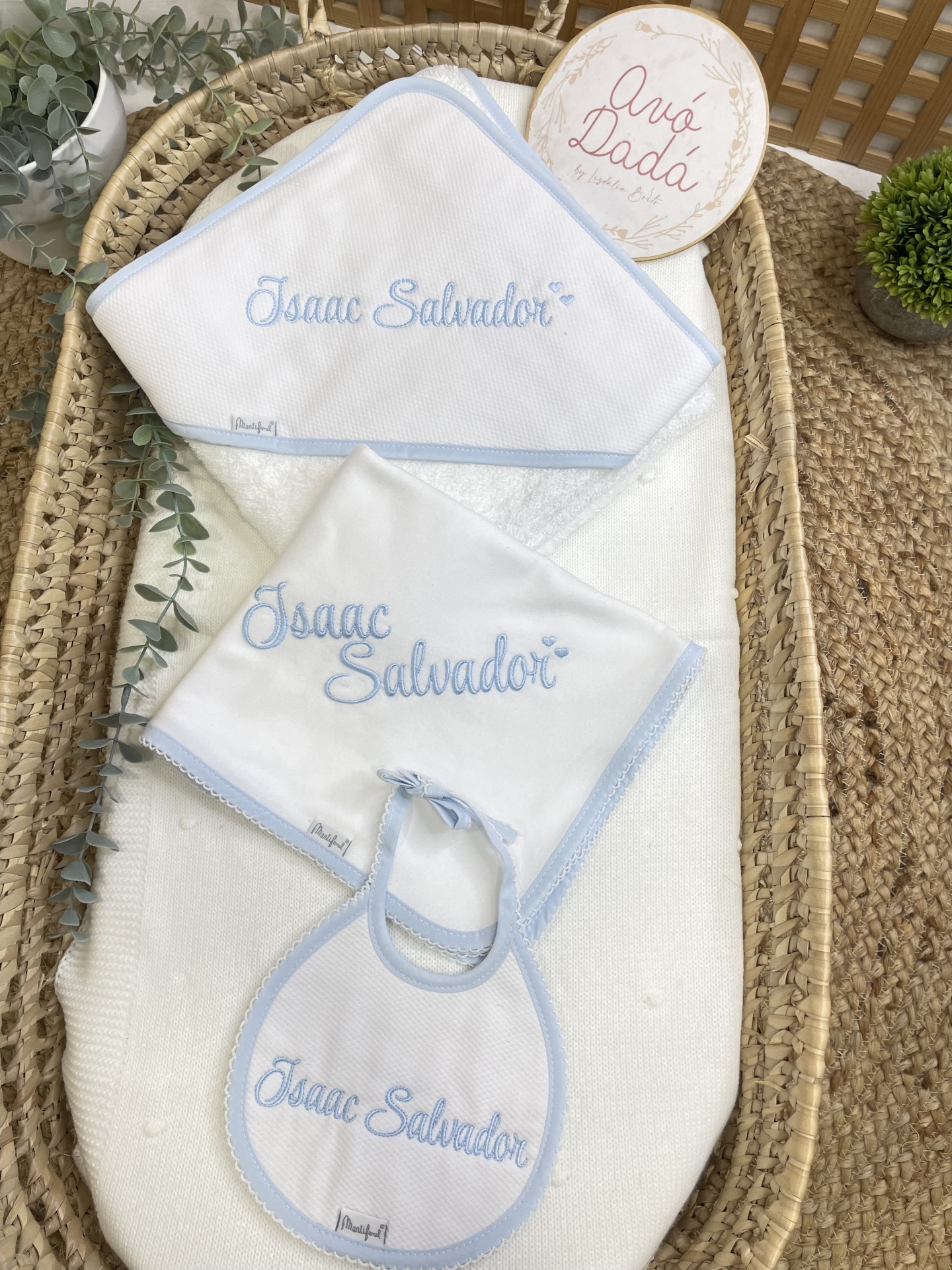 Conjunto de acessórios para bebé brancos e azuis claros com nome bordado, numa cesta de vime