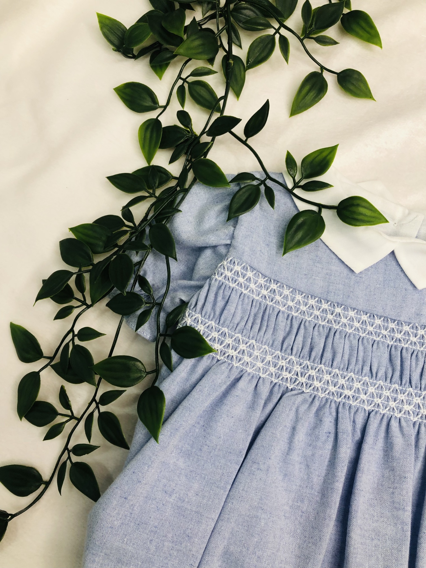 Vestido infantil azul claro com gola branca e bordado branco, com folhas verdes ao redor