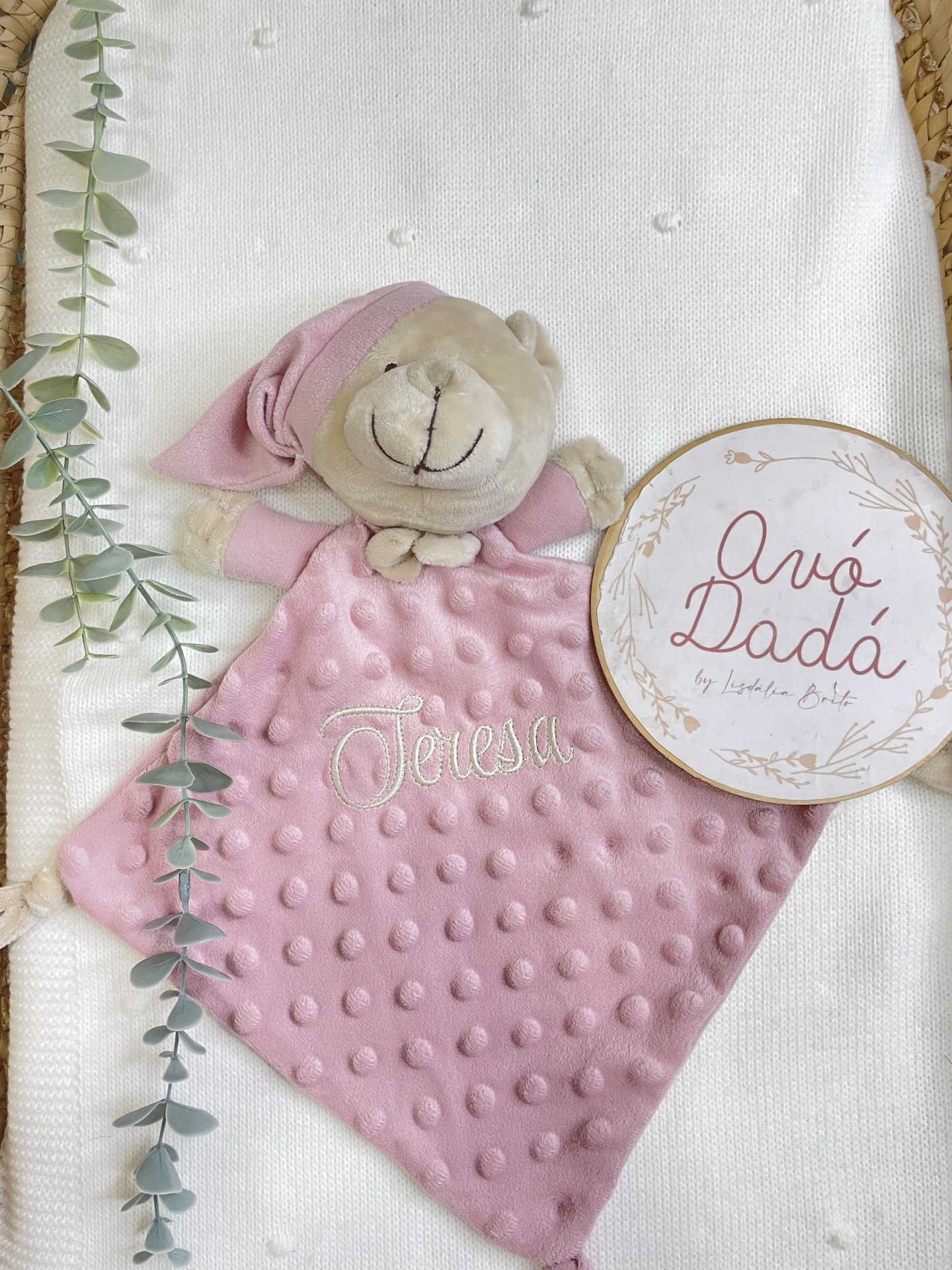 Peluche rosa com textura e nome bordado Teresa sobre tecido branco com malha, ramo verde e placa redonda com texto.