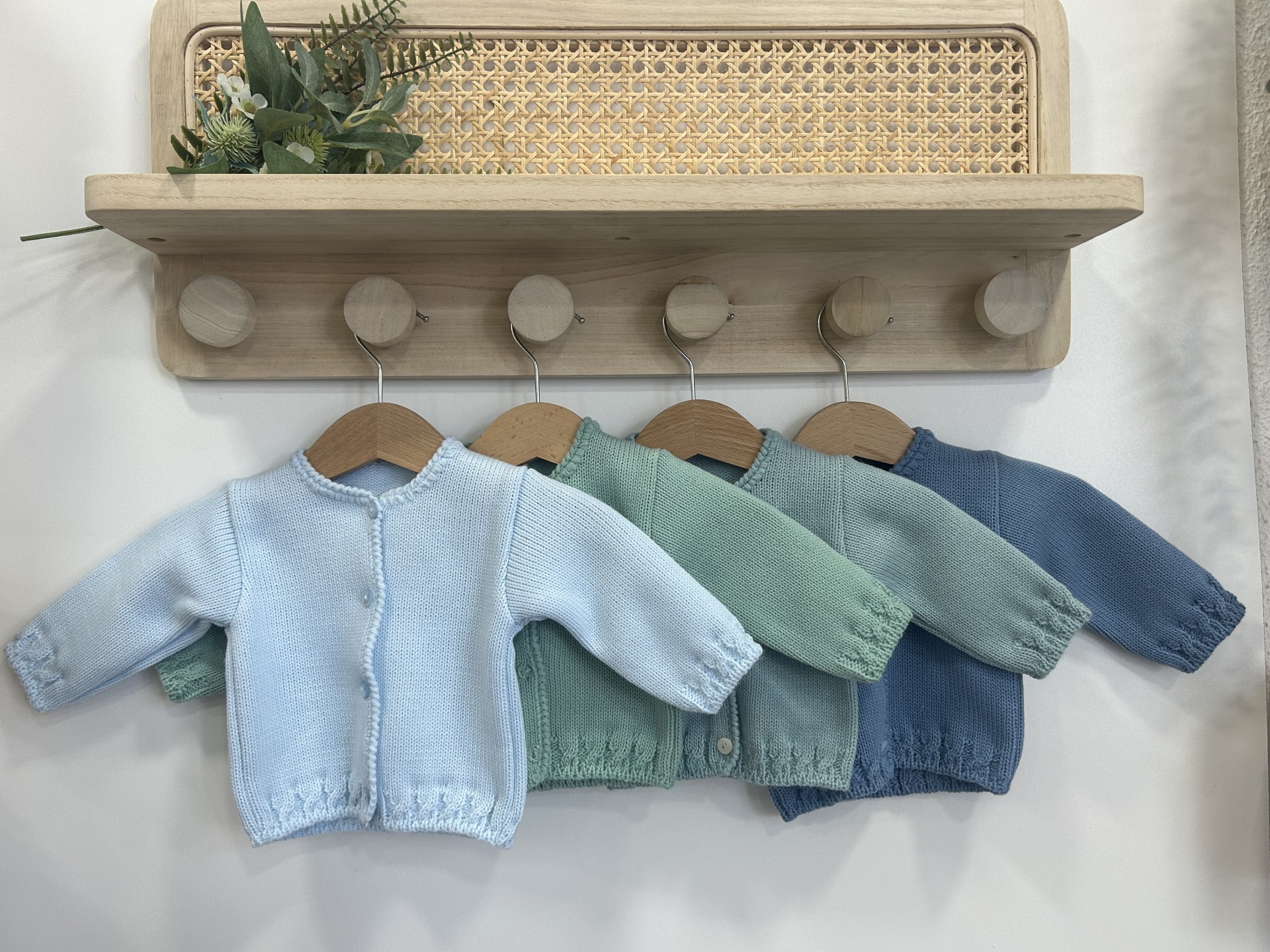 Quatro casacos de malha para bebé em cores pastel pendurados em cabides sob uma prateleira de madeira clara com detalhes em palhinha
