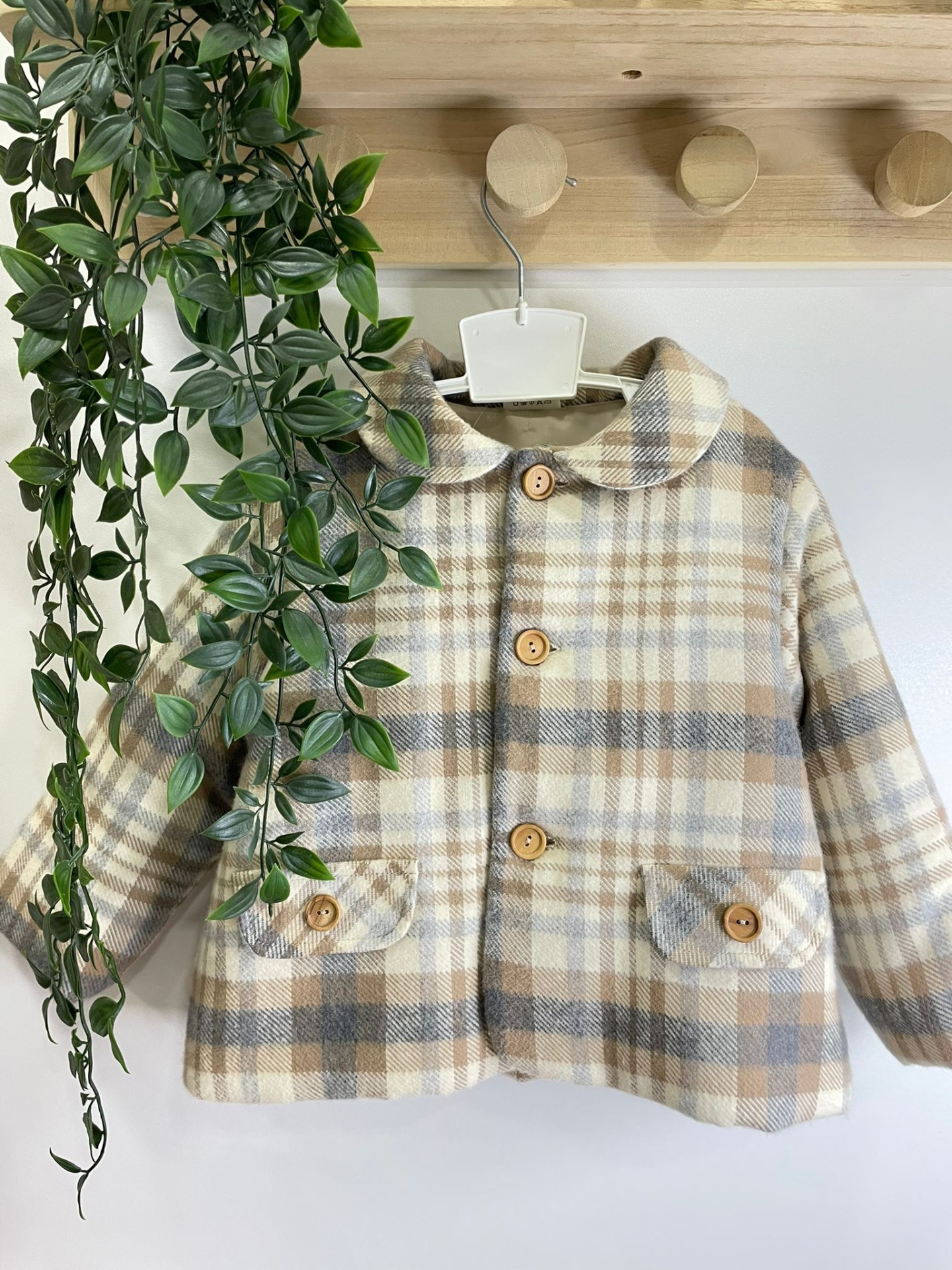 Casaco infantil xadrez bege com botões de madeira pendurado em cabideiro de madeira ao lado de planta verde