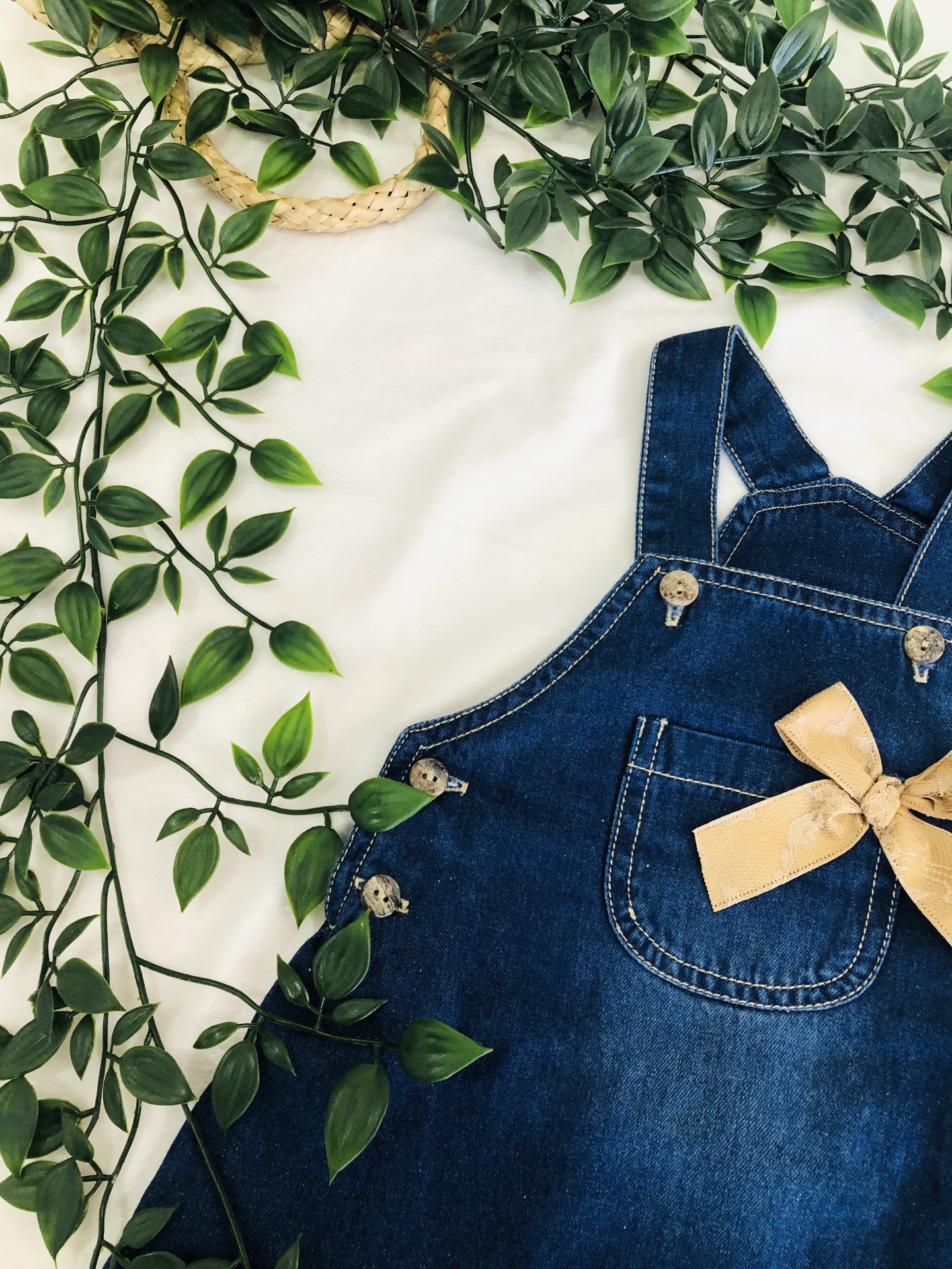 Macacão de ganga azul com alças cruzadas, laço bege e bolso, rodeado por folhas verdes