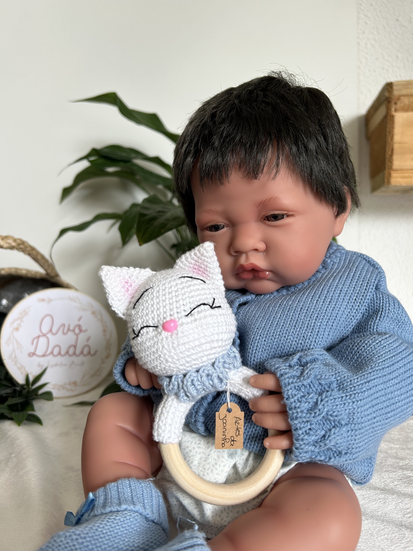 Boneco bebé com camisola azul segurando brinquedo de madeira com gato de croché