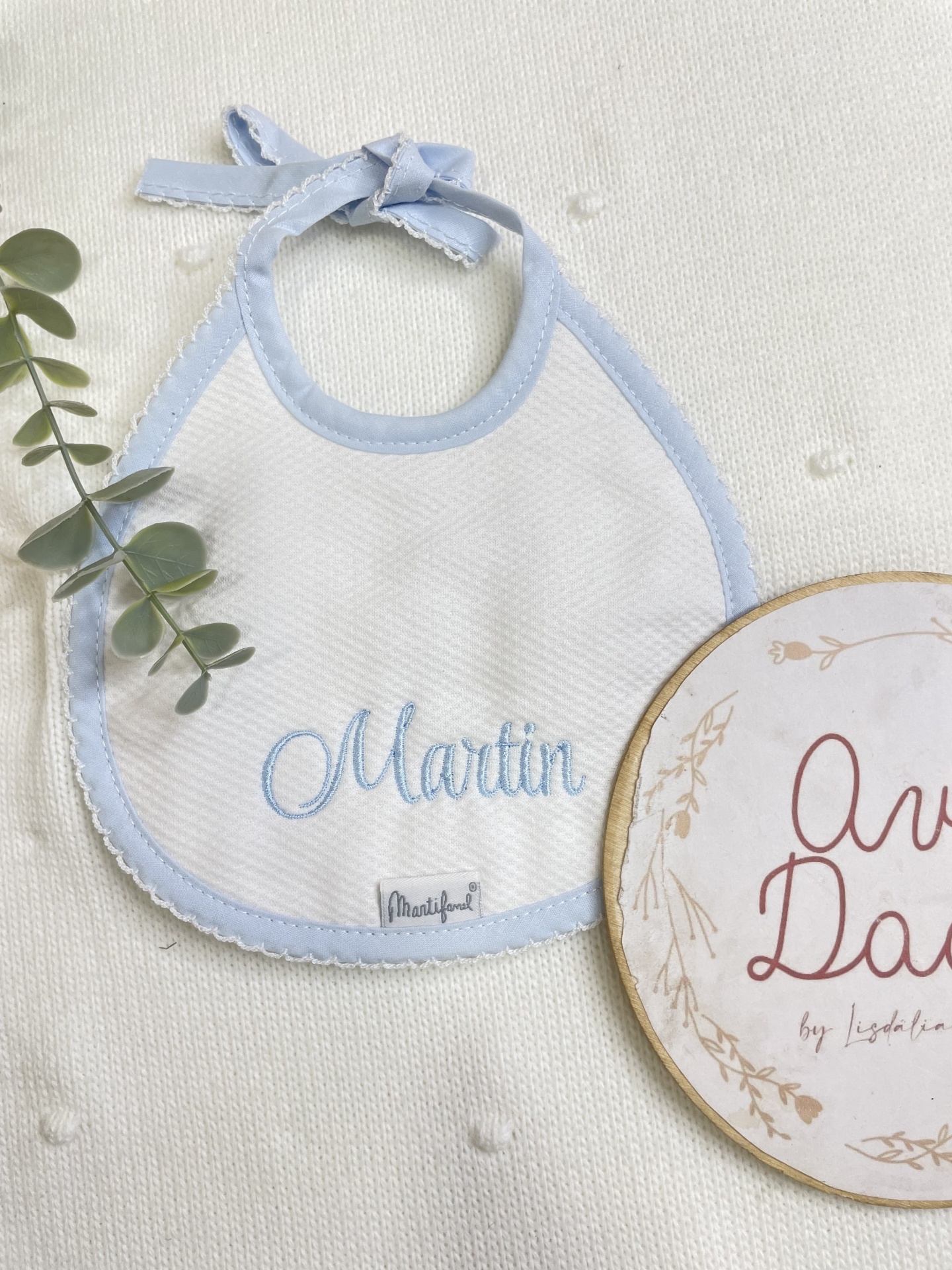 Babete branco com borda azul clara e nome Martin bordado, junto a ramo verde e placa de madeira sobre fundo creme