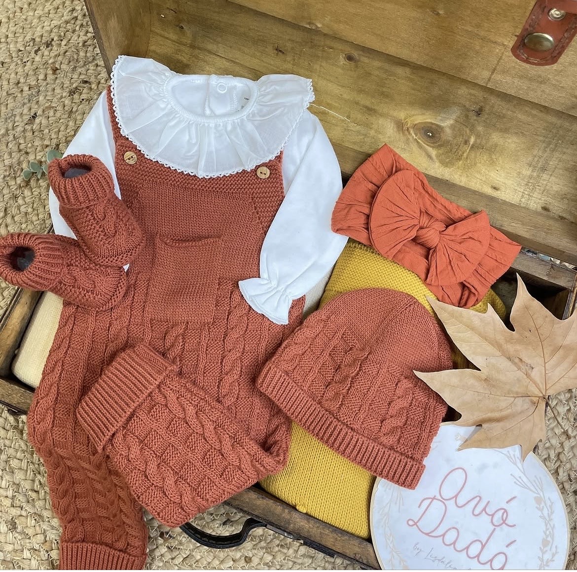 Roupa de bebé em malha terracota, camiseta branca e acessórios sobre caixa de madeira