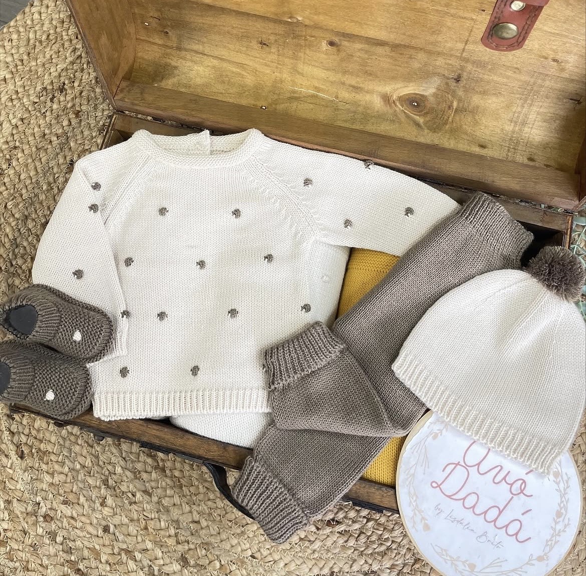 Conjunto de roupa de bebê em malha com camisola, calças, gorro e sapatinhos numa mala de madeira.