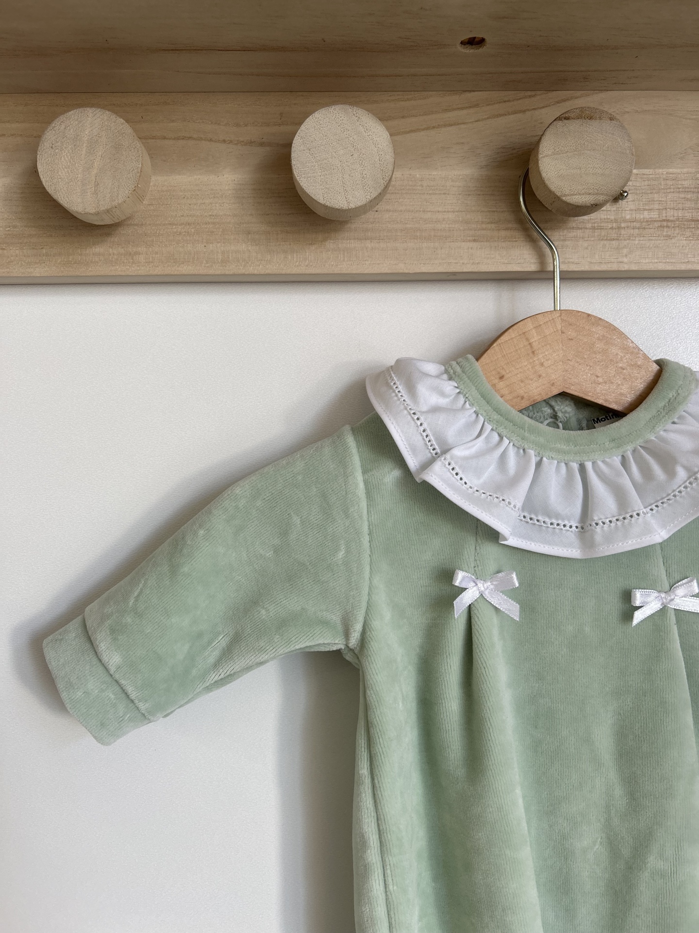 Roupa de bebé verde com gola branca de babados e dois laços brancos pendurada num cabide de madeira
