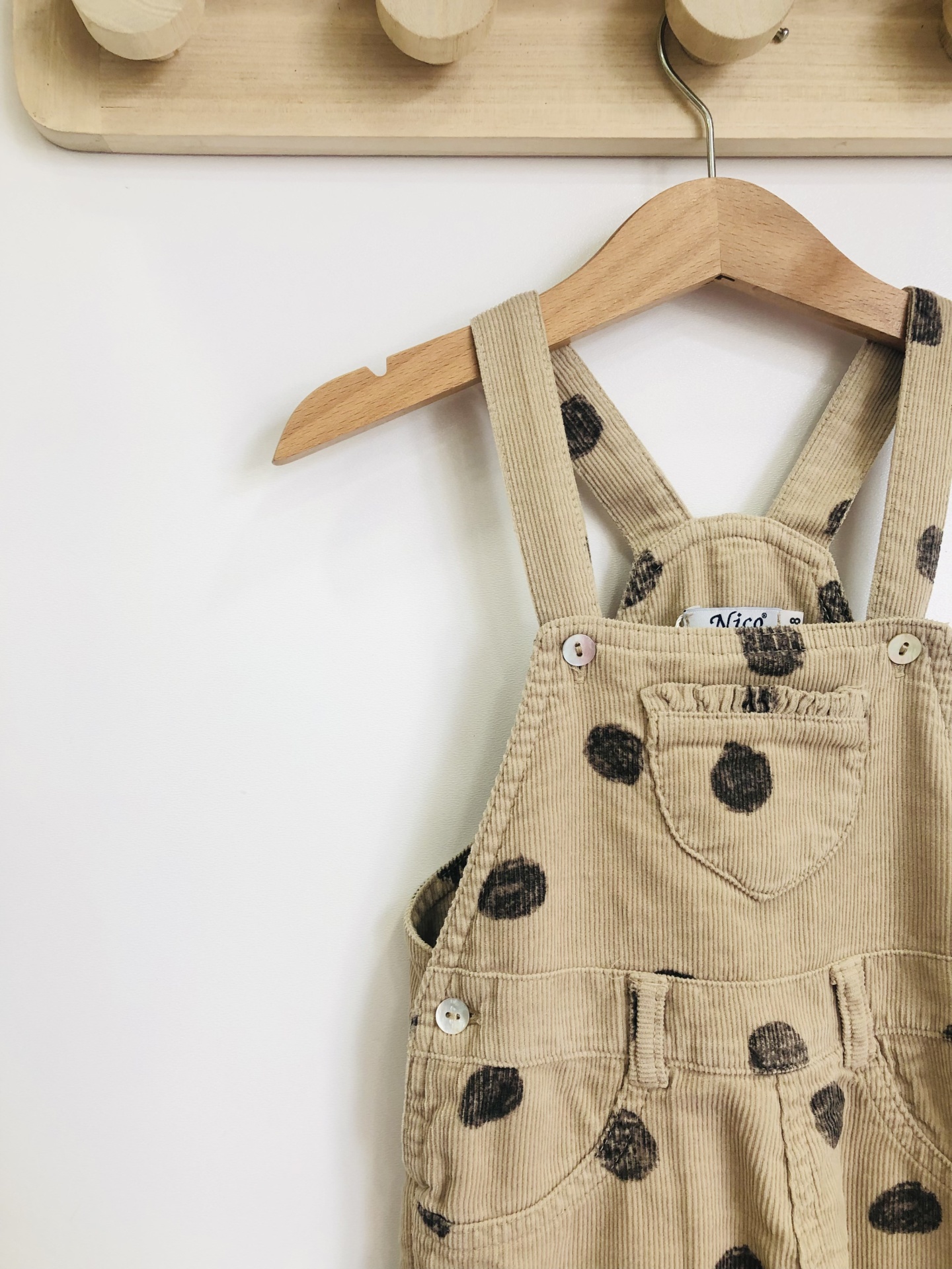 Jardineiras de criança bege com bolinhas pretas penduradas em cabide de madeira