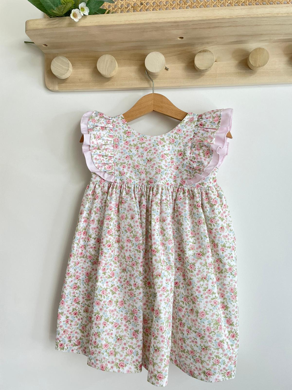 Vestido infantil floral branco com folhos lilases pendurado em cabide numa parede