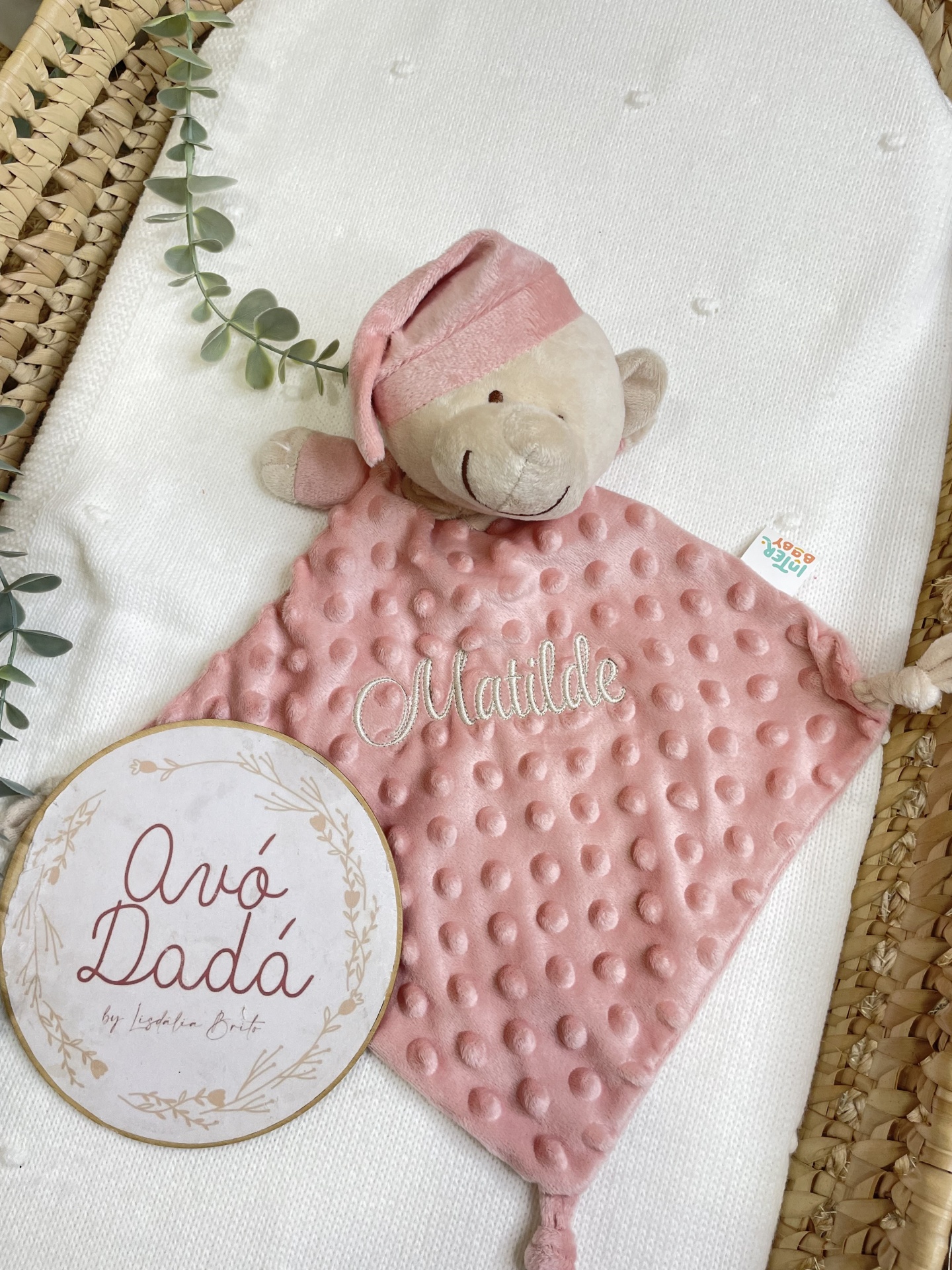 Manta rosa com ursinho de peluche e bordado Matilde, quadro decorativo com texto Avó Dadá, sobre fundo branco e cesta de palha.