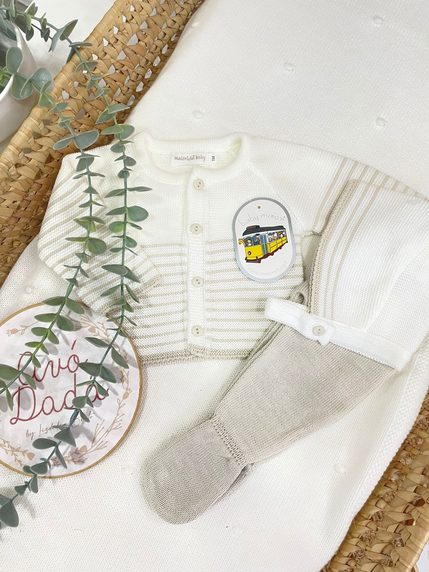 Conjunto de roupa de bebé branca e bege com etiqueta baby maiors num cesto de palha com planta artificial
