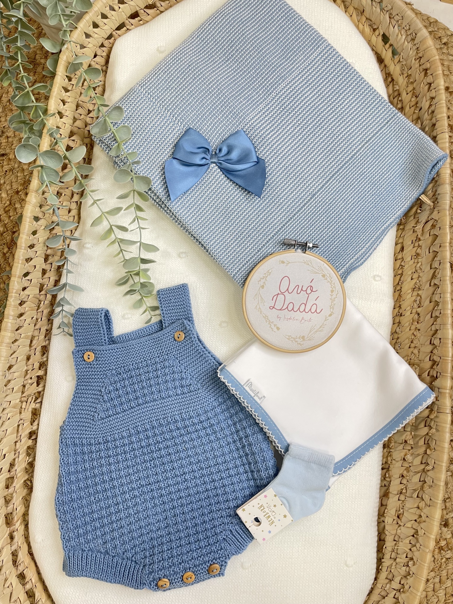 Conjunto de roupas e acessórios para bebé em azul e branco sobre uma cesta de vime.