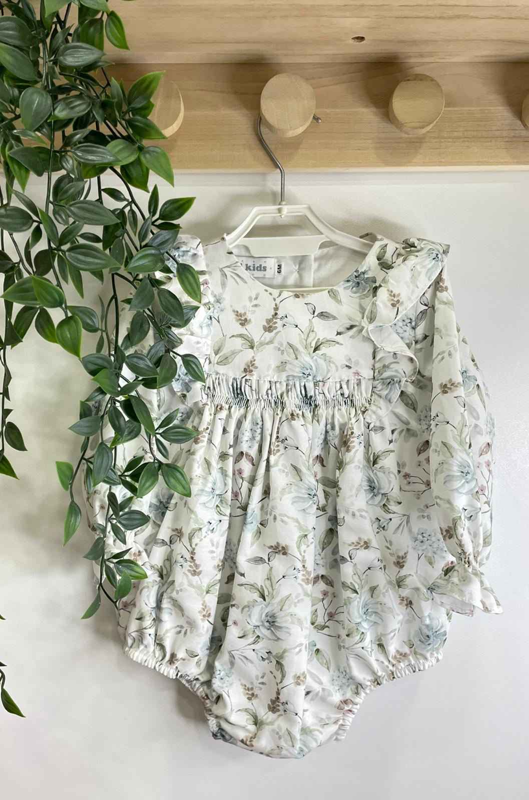 Body para bebé com padrão floral em tons suaves, pendurado num cabide branco junto a uma planta verde e suporte de madeira