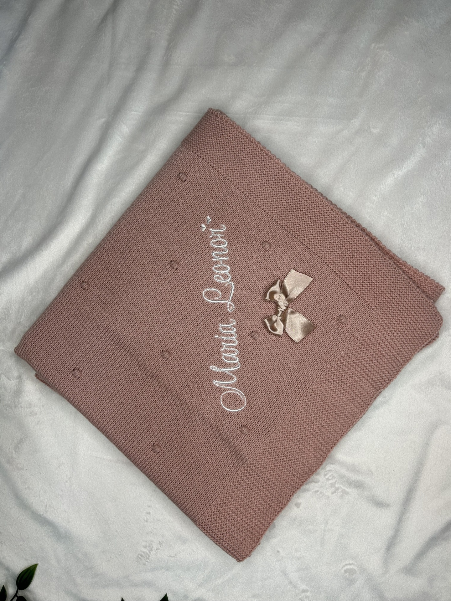 Manta rosa com laço bege e texto bordado 'Manta Lenore' sobre superfície branca