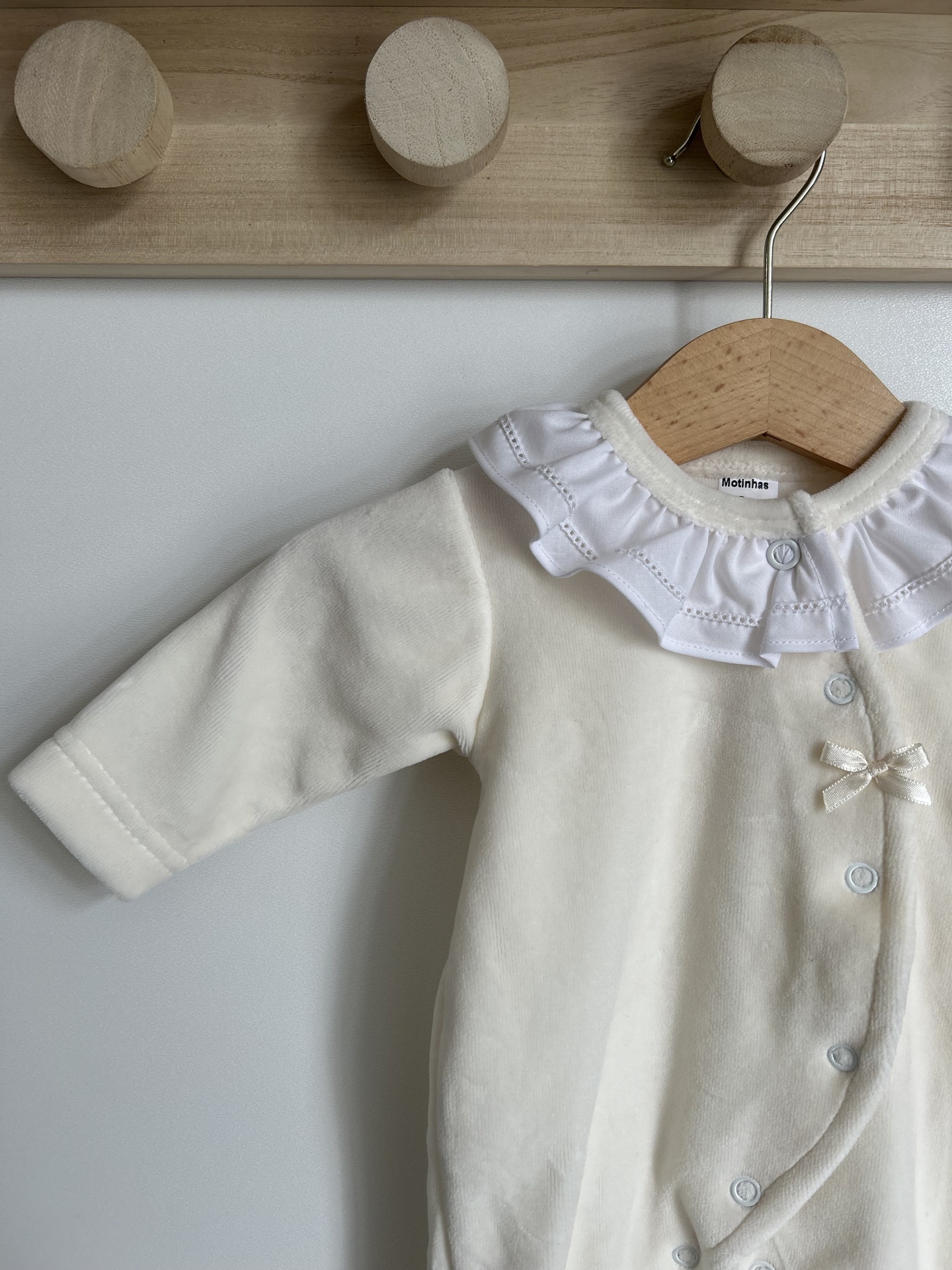macacão de bebé creme com gola branca e botões azuis pendurado em cabide de madeira