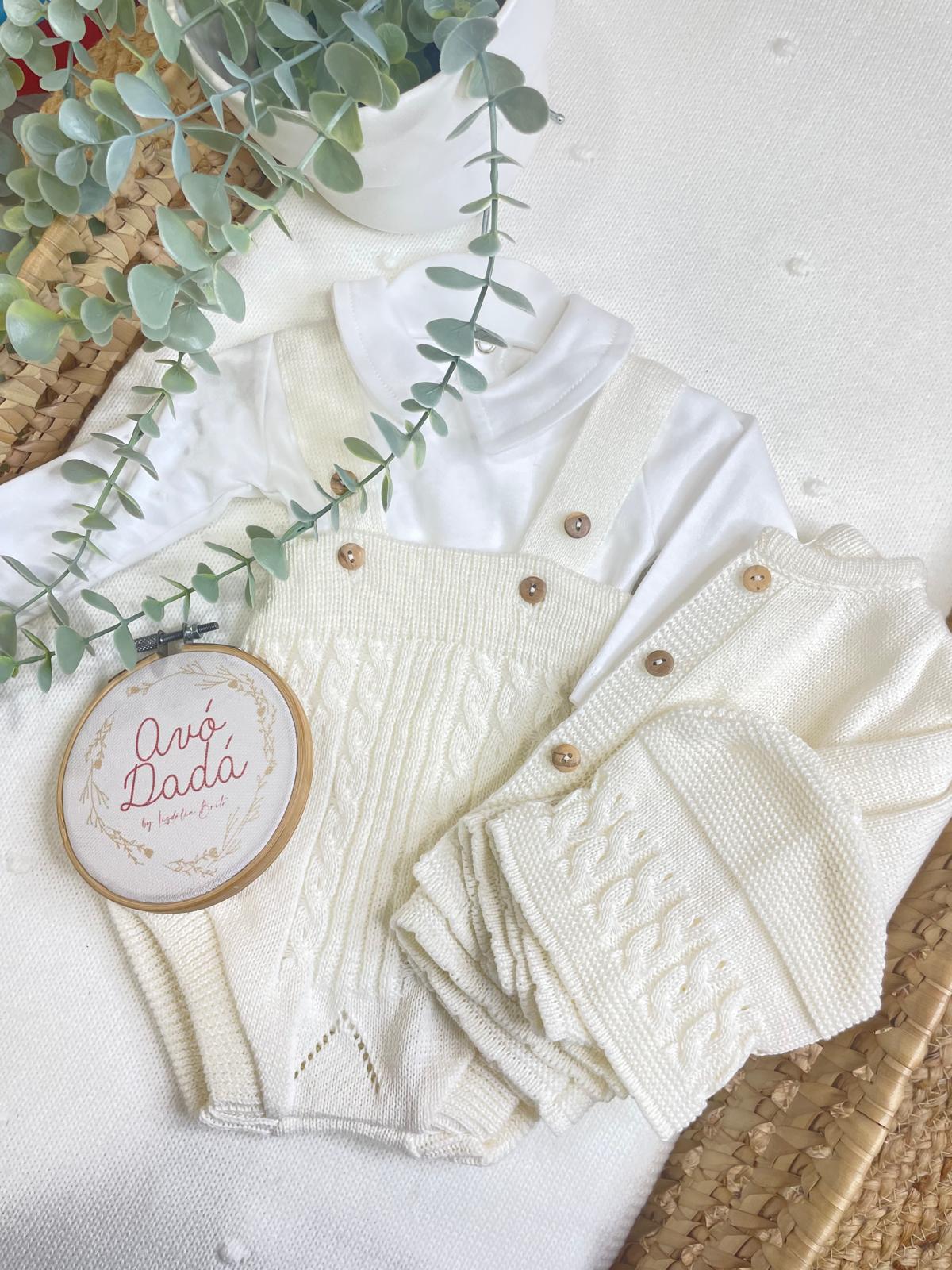 Conjunto de roupa de bebé creme com body branco, jardineira e casaco tricotados, planta verde e placa decorativa com texto Avó Dadá.