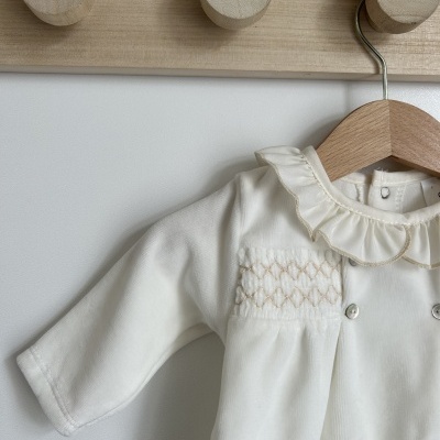 Macacão de bebé branco com gola folhos e bordados pendurado numa parede