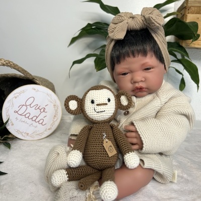 Boneca bebé com roupa de malha e faixa na cabeça, segurando macaco de croché, planta e caixa ao fundo