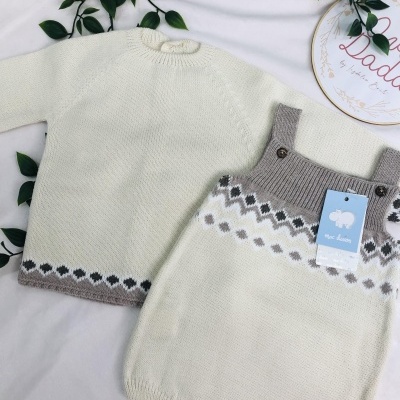 Conjunto de roupa de bebé em malha branca com padrão cinzento e bege, incluindo camisola e jardineiras, sobre tecido branco com folhas verdes.