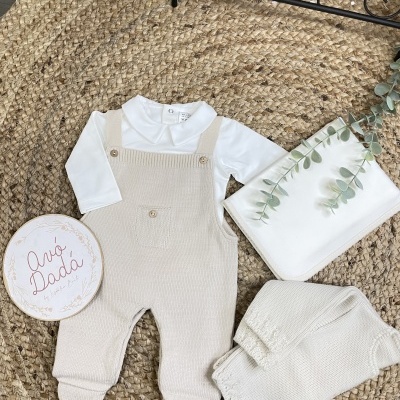 Conjunto de roupa de bebé com body branco e jardineira de malha bege sobre tapete de fibras naturais